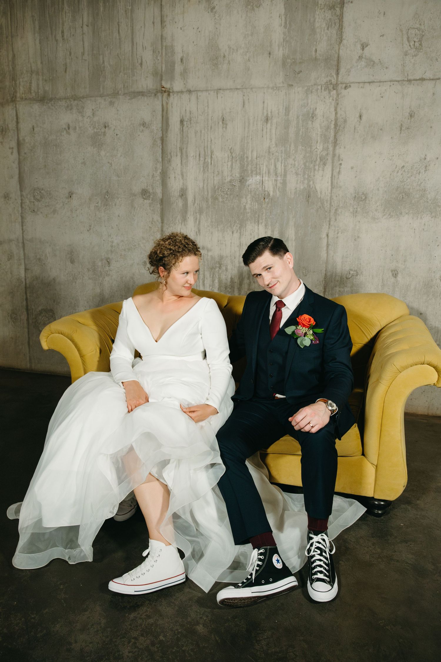Wedding couple lounges playfully on yellow couch wearing formal attire and casual sneakers in industrial setting.