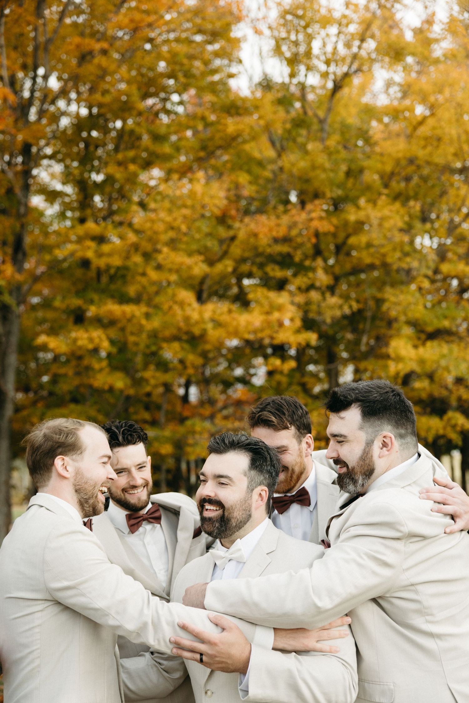 A group of groomsmen in beige suits share a joyful moment together against a backdrop of golden autumn trees.