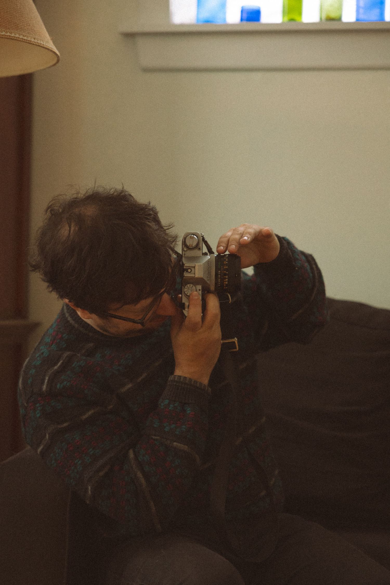 Person sitting on sofa photographing with a vintage film camera in a cozy dim room.