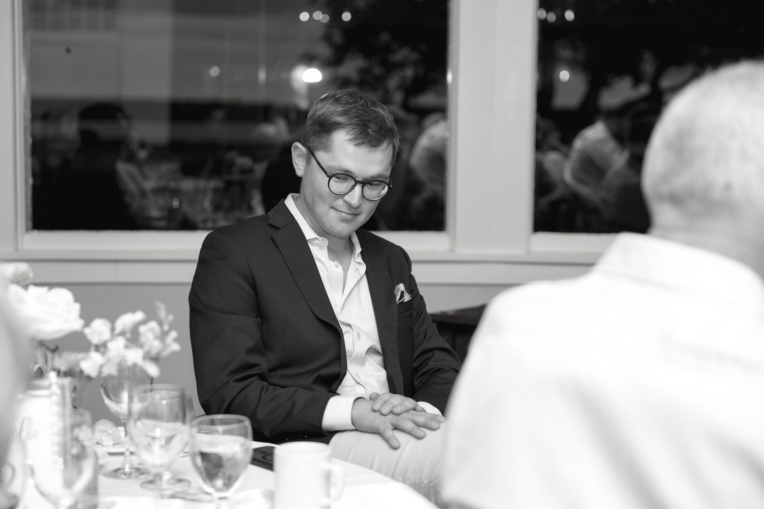 Man in dark blazer looking down at hands during wedding reception dinner in black and white photo.