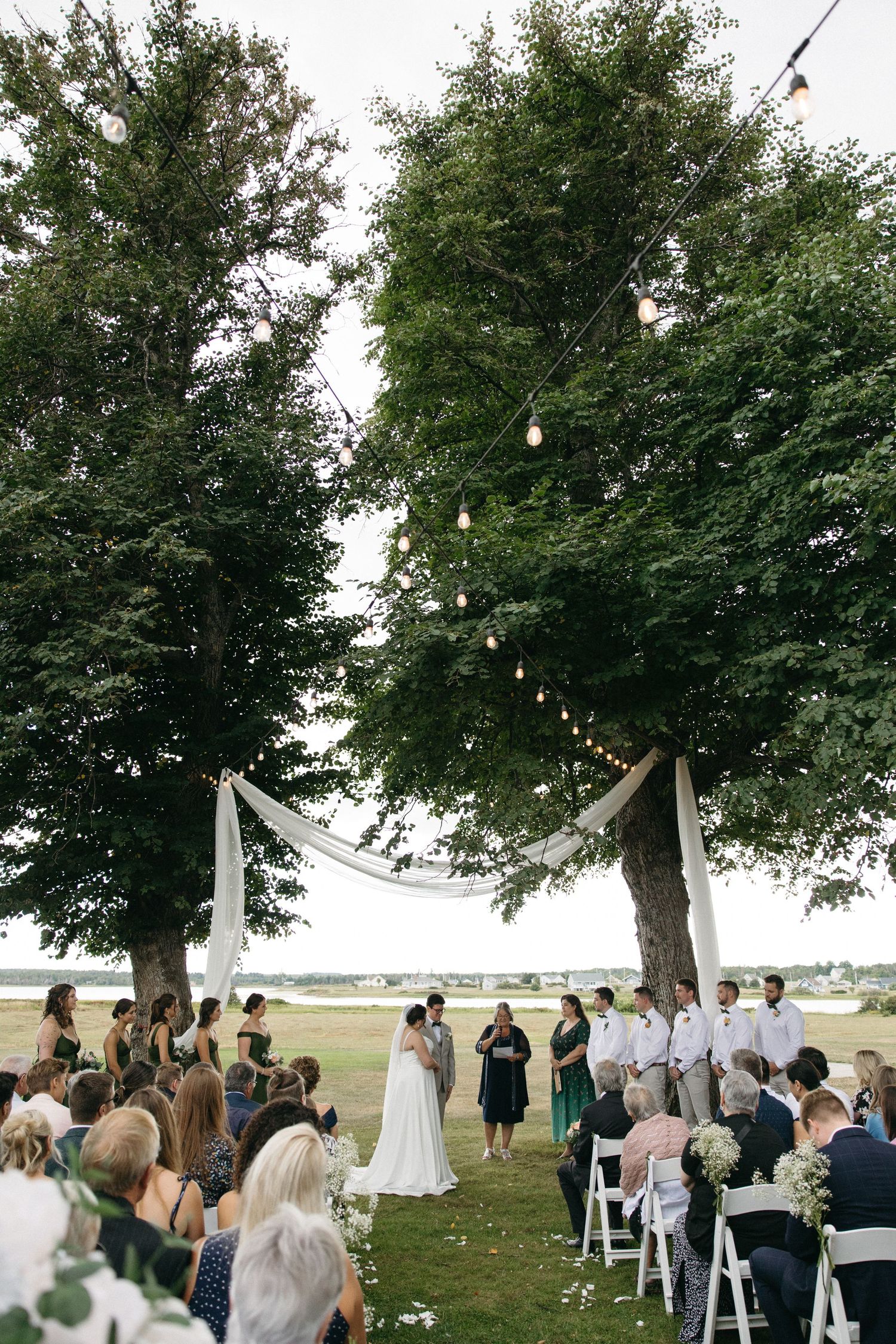 Outdoor wedding ceremony under two large trees with string lights and white draping near a waterfront.