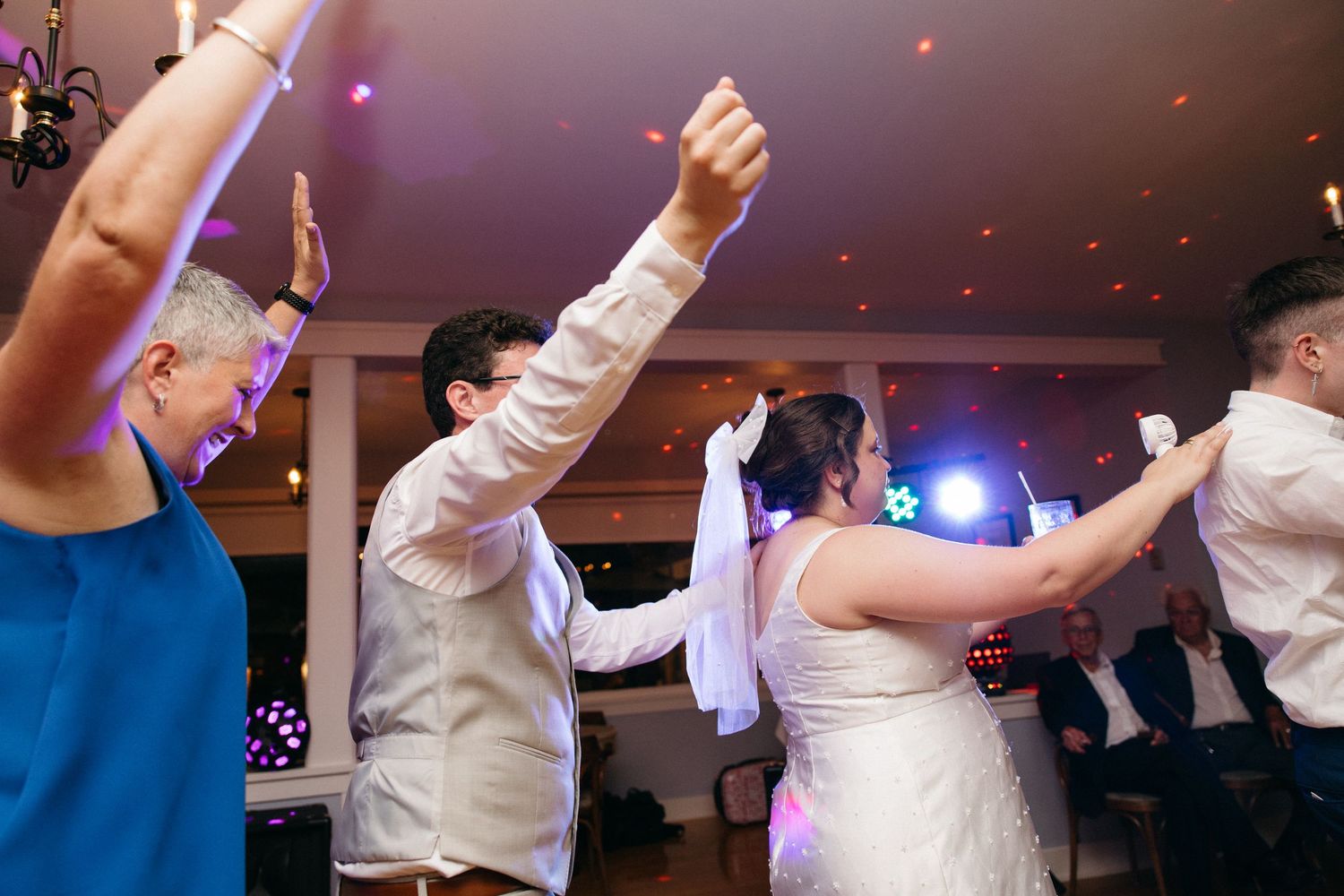 Wedding guests and couple dancing joyfully at reception with colorful lights.