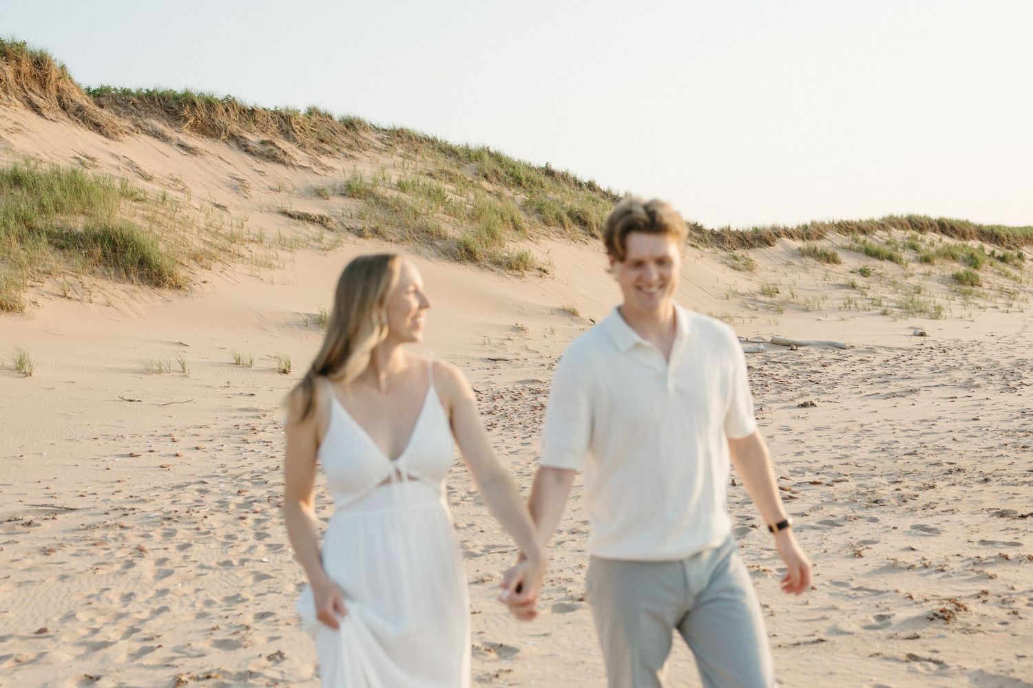 Couple holding hands walking on a sandy beach, woman in white dress and man in casual attire.