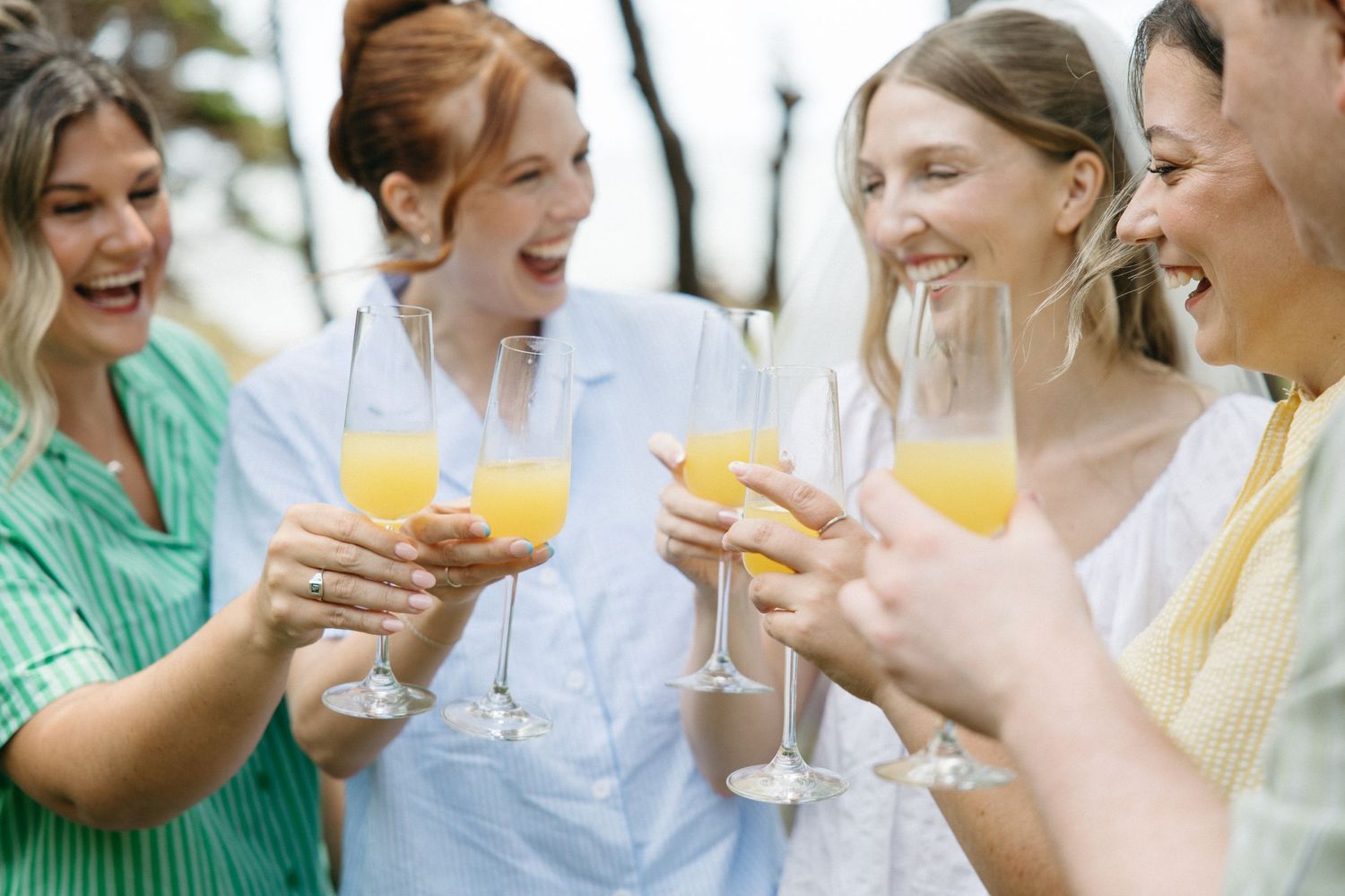 Four women smiling and toasting with mimosas at an outdoor brunch celebration.