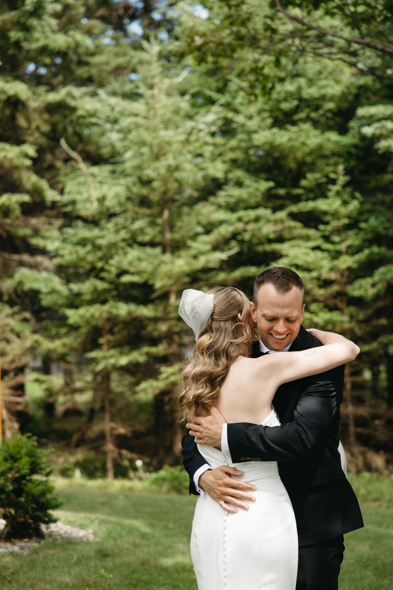 Bride and groom share a joyful first look hug outdoors surrounded by lush green pine trees.