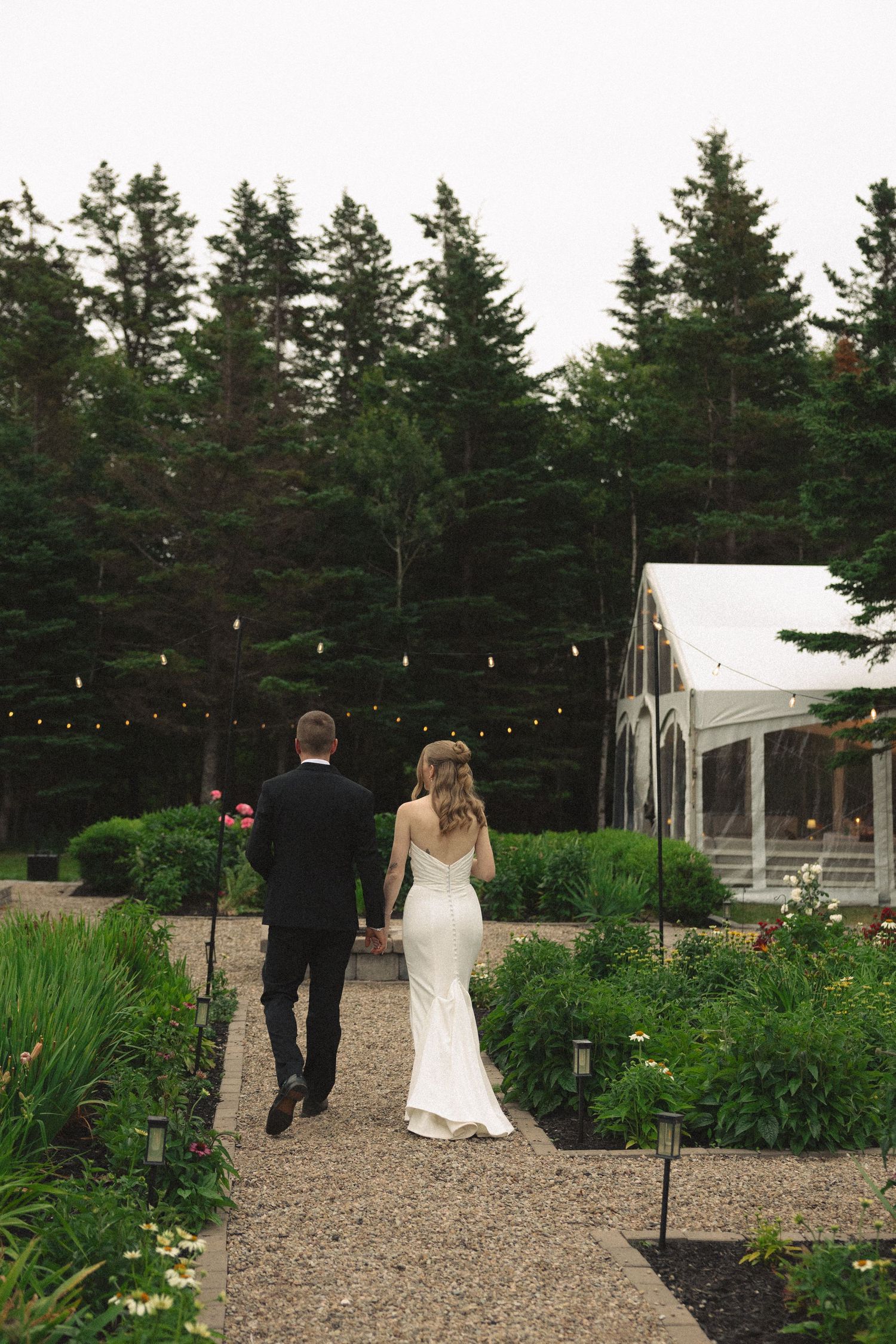 Bride and groom walk hand in hand toward lit reception tent through lush garden path.