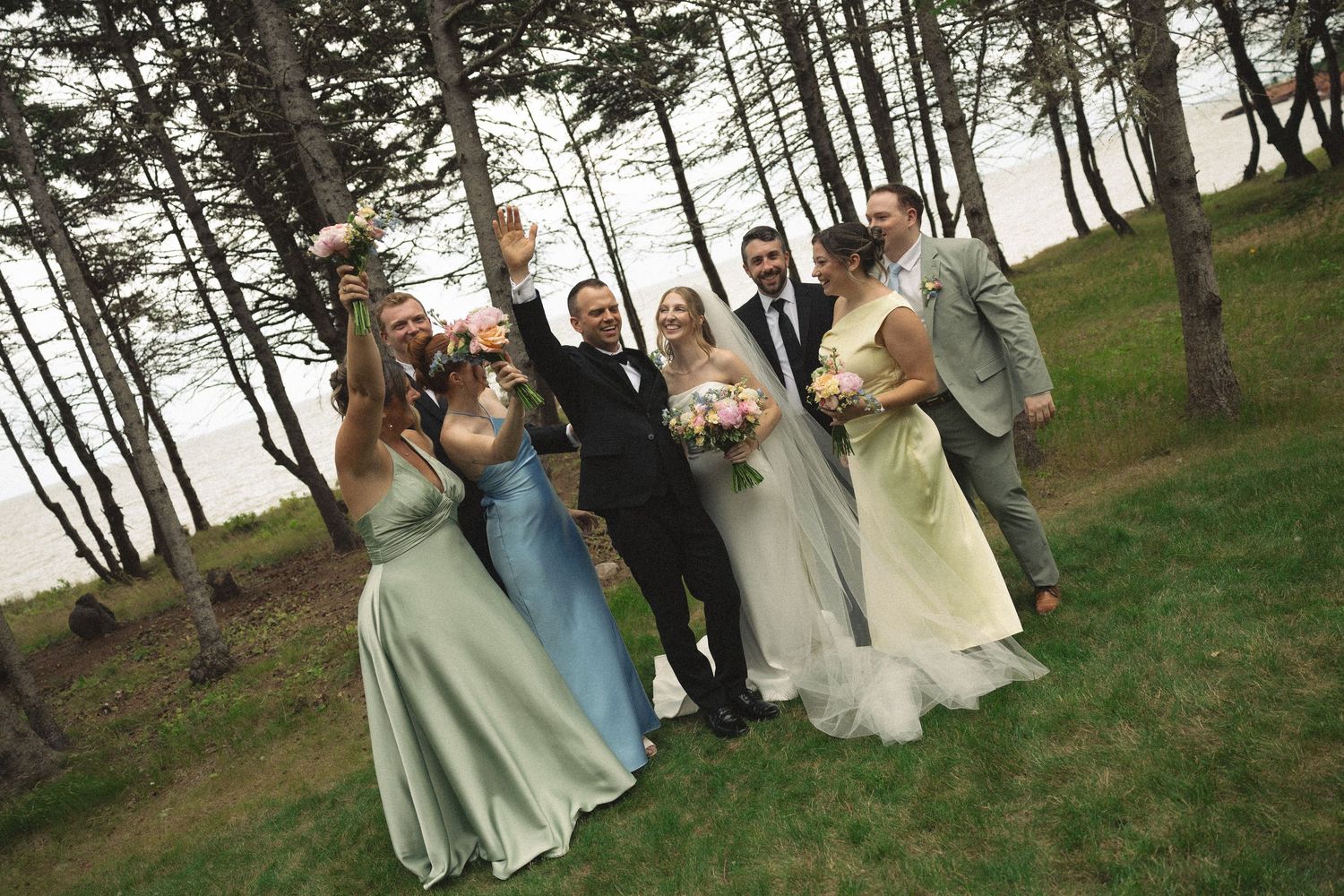Wedding party celebrates together among tall pine trees near a waterfront, laughing and raising bouquets.