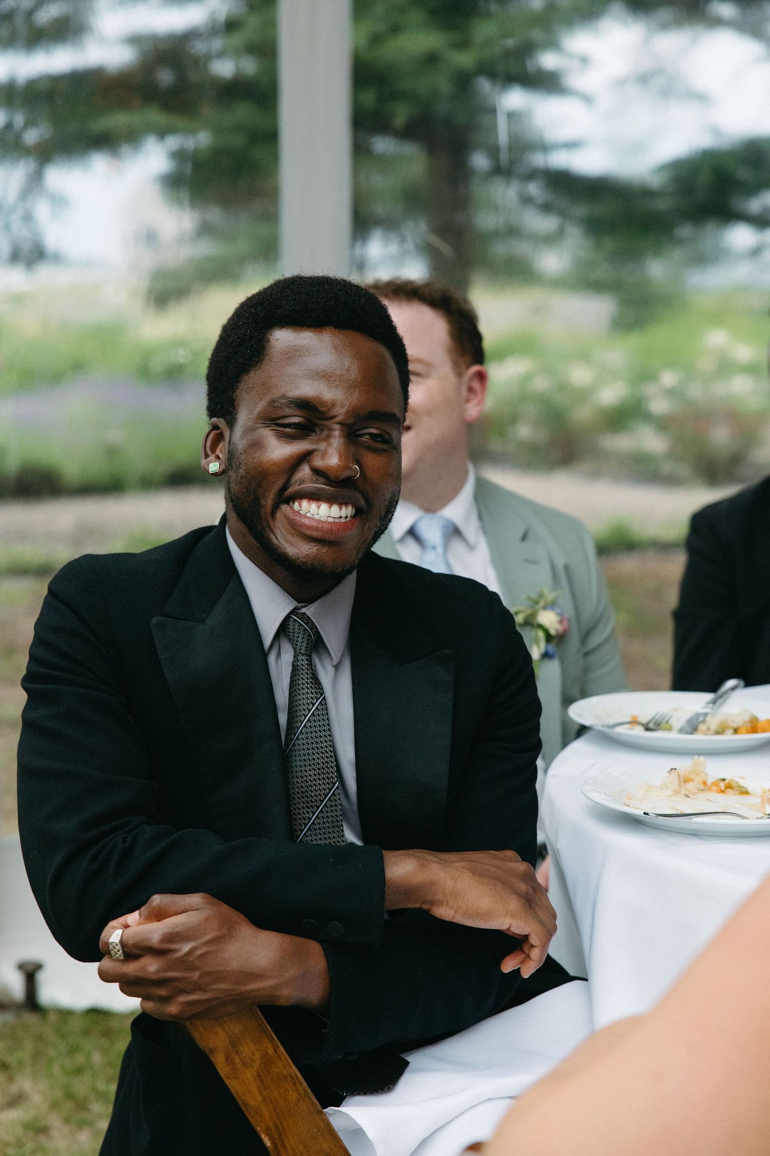 Smiling man in black suit and tie laughs joyfully while seated at outdoor wedding reception tent.
