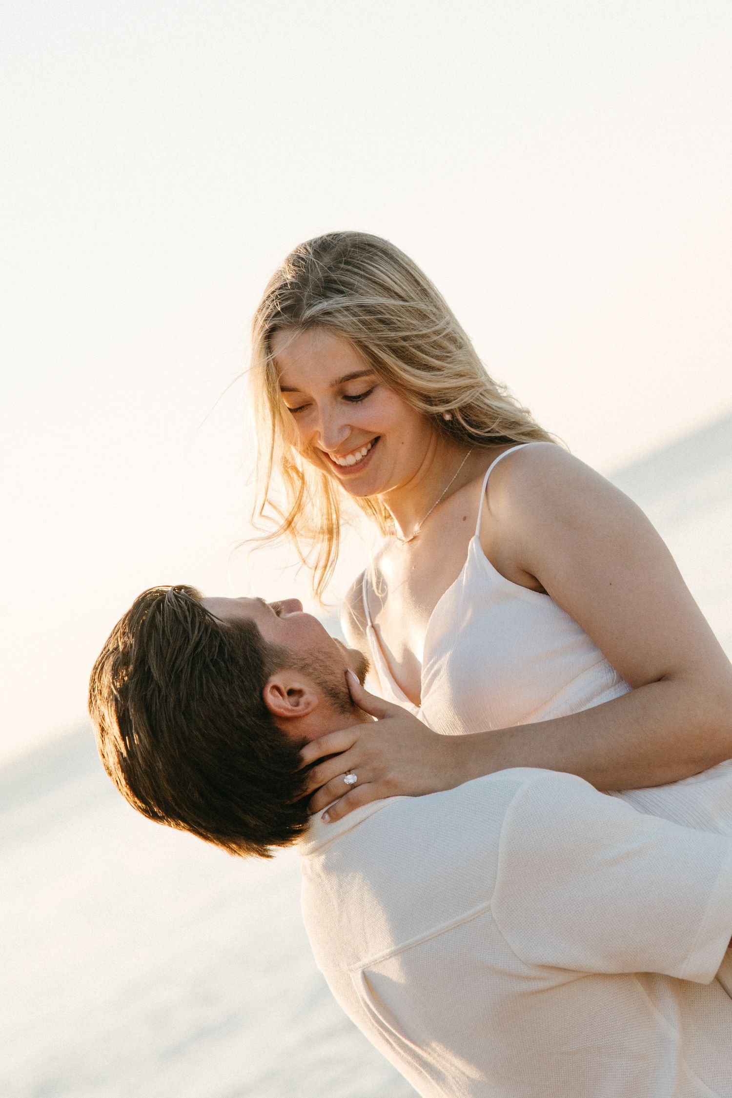 Man lifting smiling blonde woman in white dress on beach at golden hour.