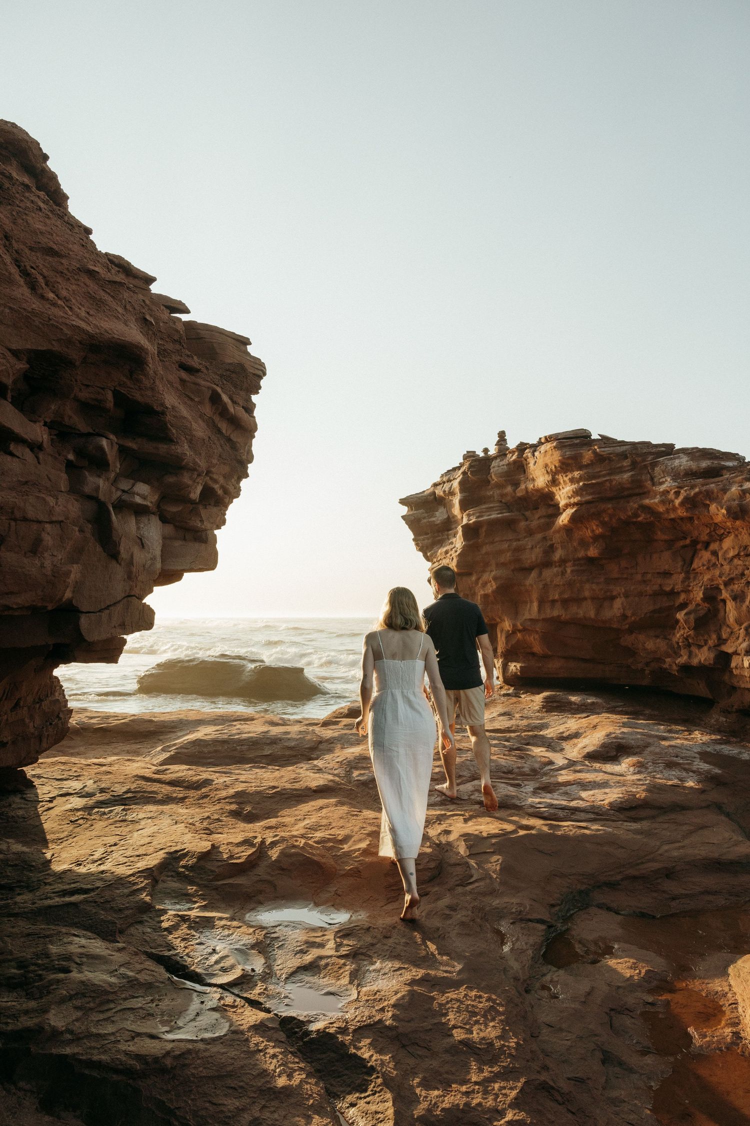 Two people explore dramatic red rock formations by the ocean during sunset.