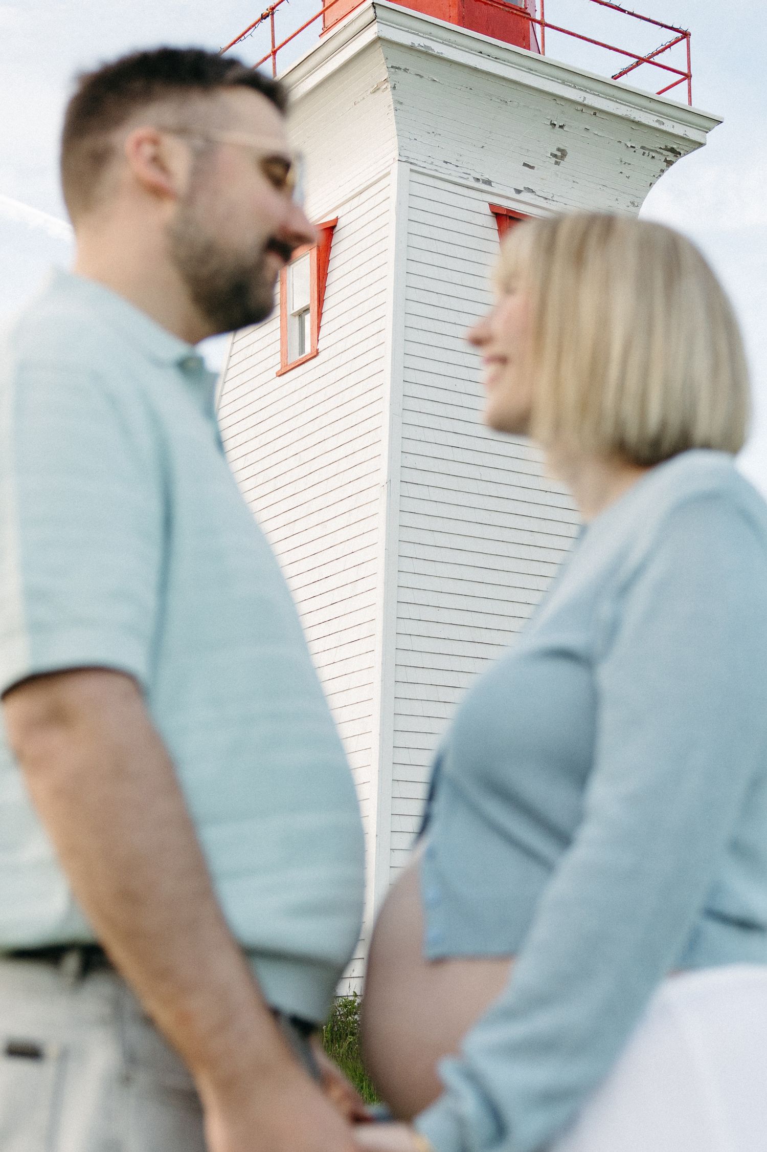 Expecting couple holds hands and smiles at each other in front of a white and red lighthouse.