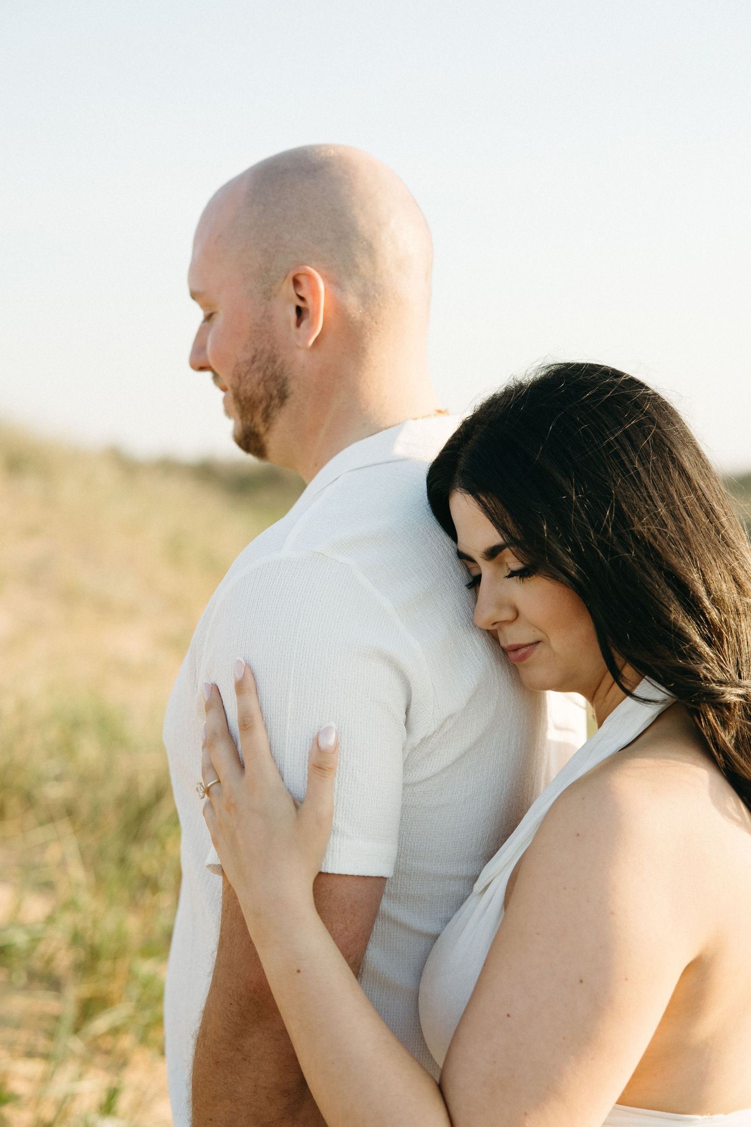 A couple embraces outdoors, woman with dark hair rests her head on a bald mans back in a sunny field.