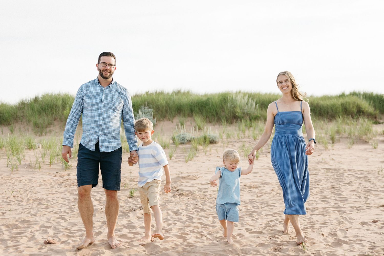 Happy family of four holding hands while walking on a sandy beach with dune grass behind them.