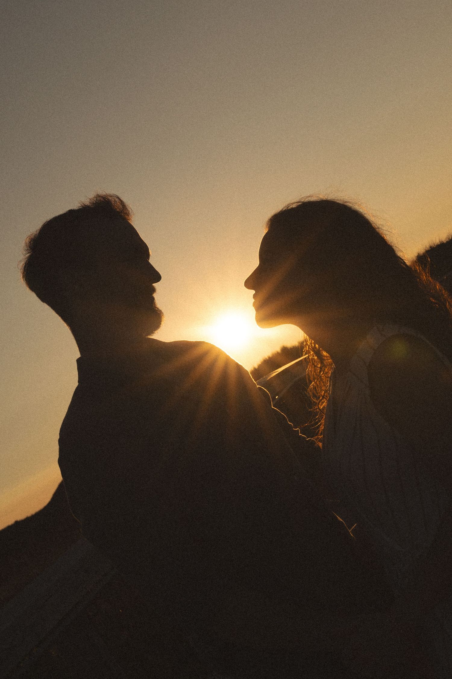 Romantic couple silhouette at golden sunset, sun rays glowing between them in an intimate embrace outdoors.