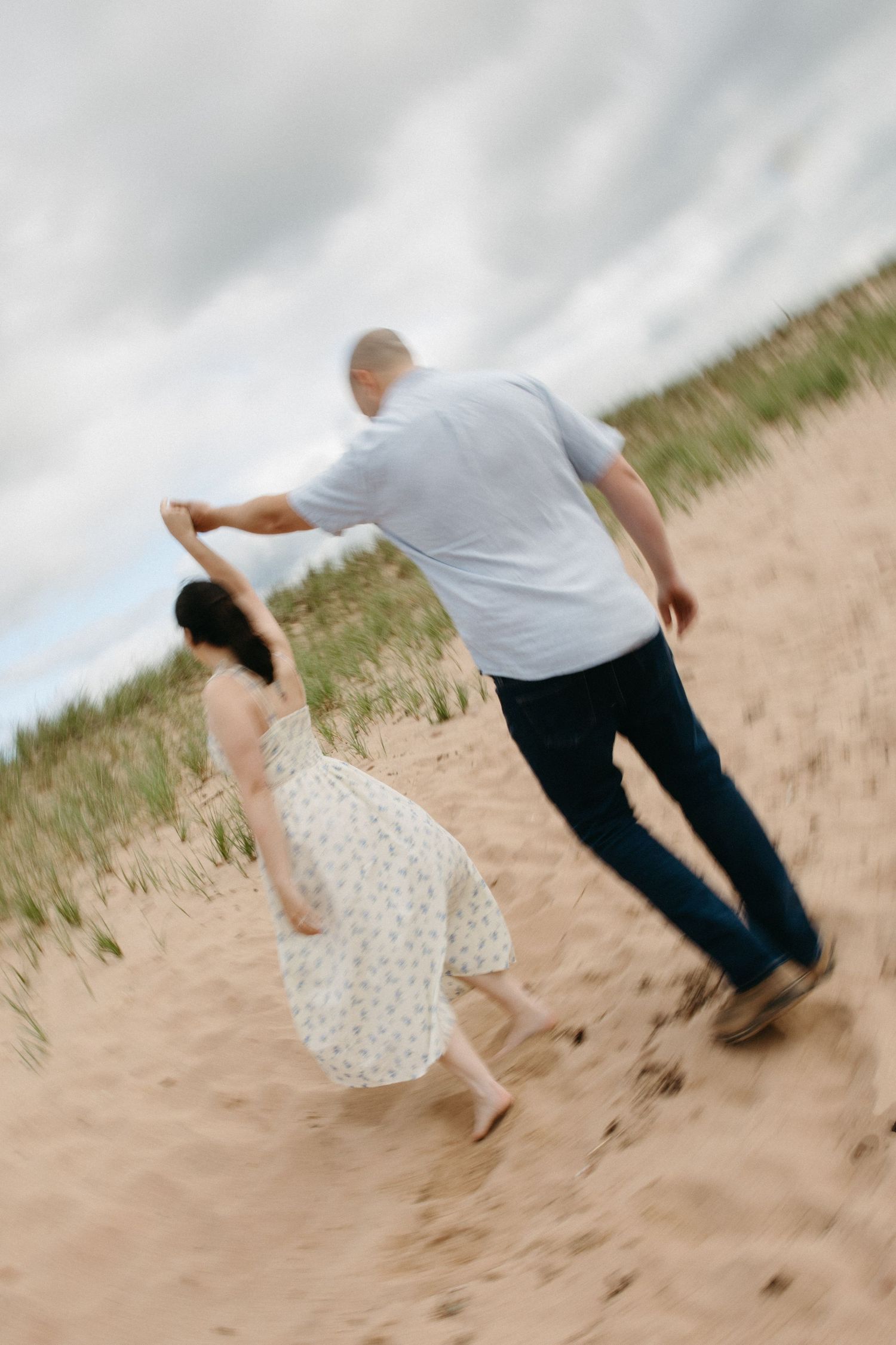 Couple holding hands walking up a sandy dune, woman in floral dress, motion blur effect on cloudy day.