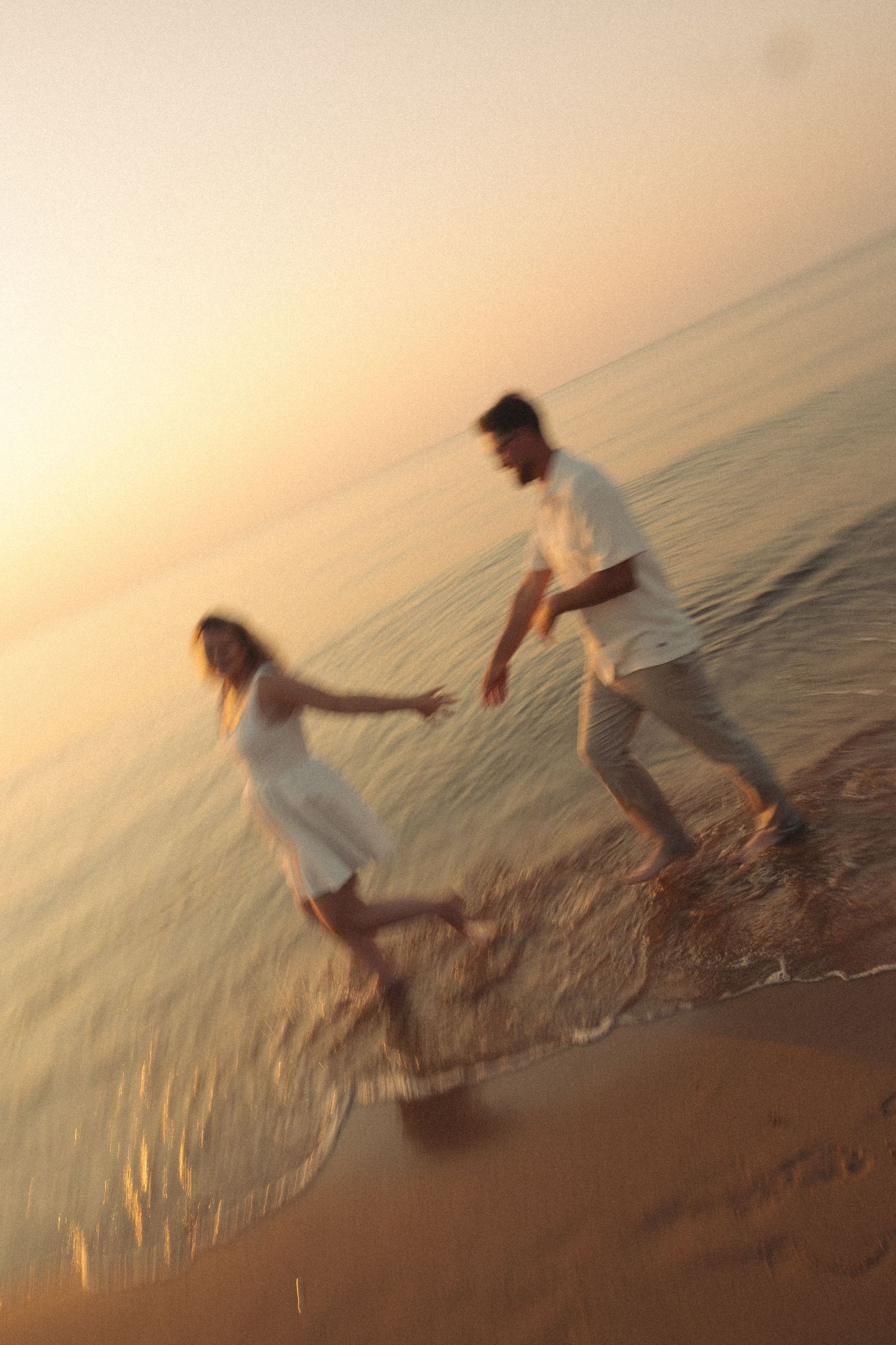 Couple holding hands and splashing in ocean waves at sunset, wearing white outfits on a sandy beach.