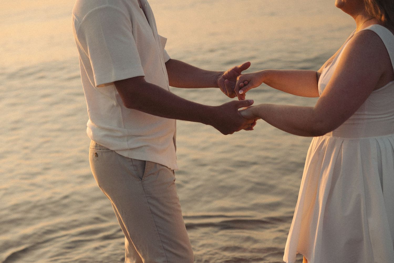 Couple holding hands on a beach at sunset, wearing white and beige outfits near calm ocean waters.