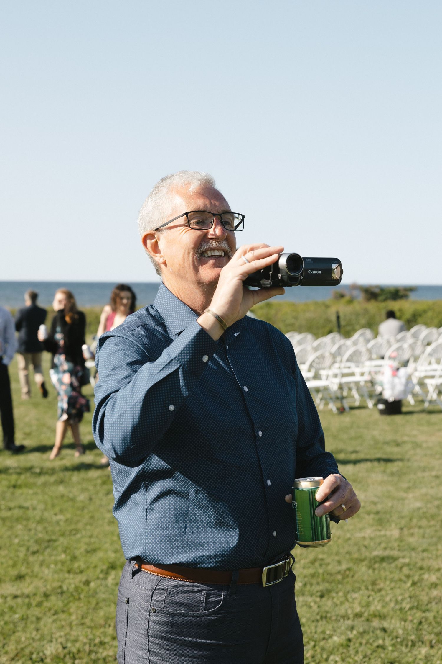 A videographer wearing glasses and a blue shirt captures footage at an outdoor event with white chairs.
