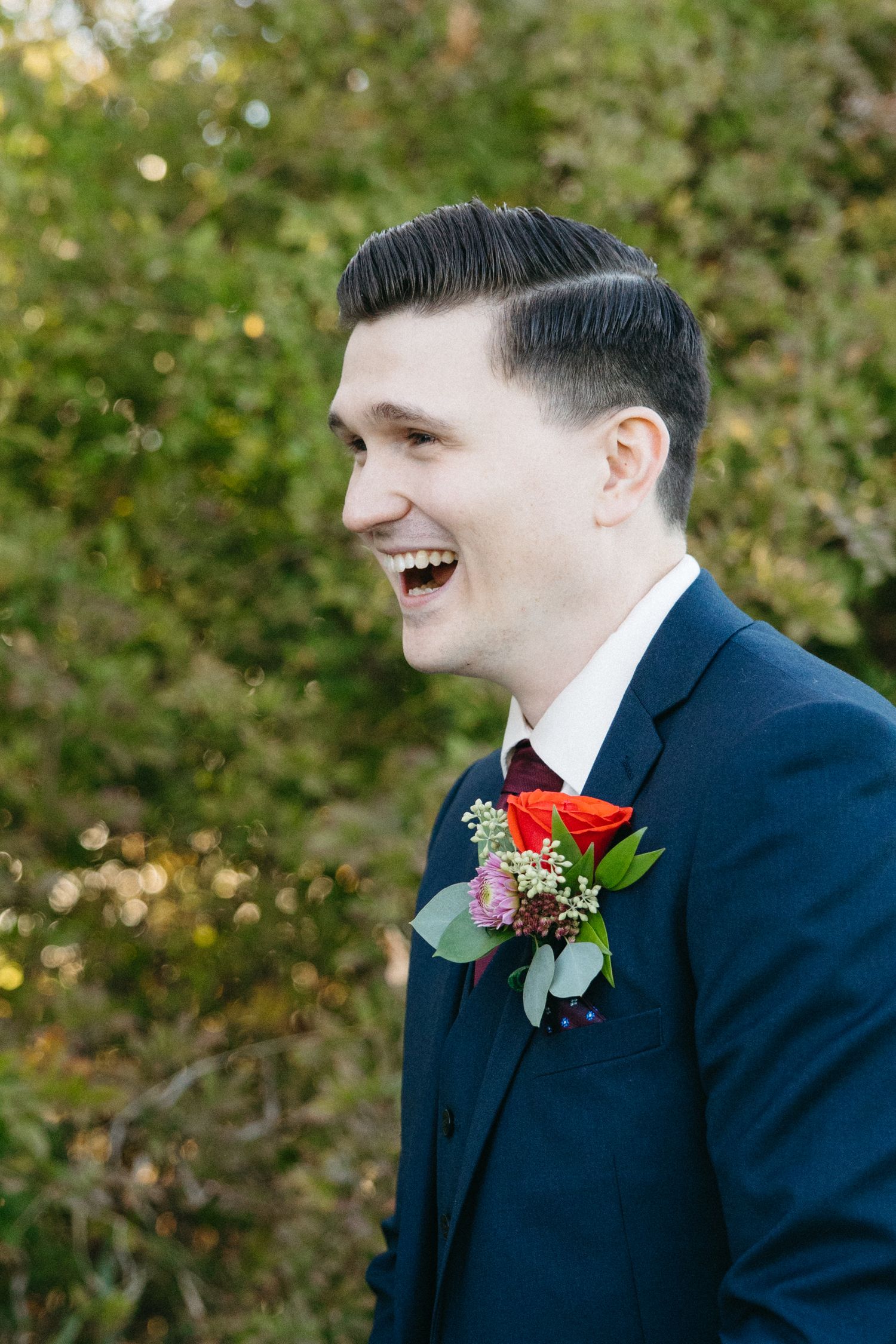 A red boutonniere adorns a navy suit jacket as the wearer smiles warmly.