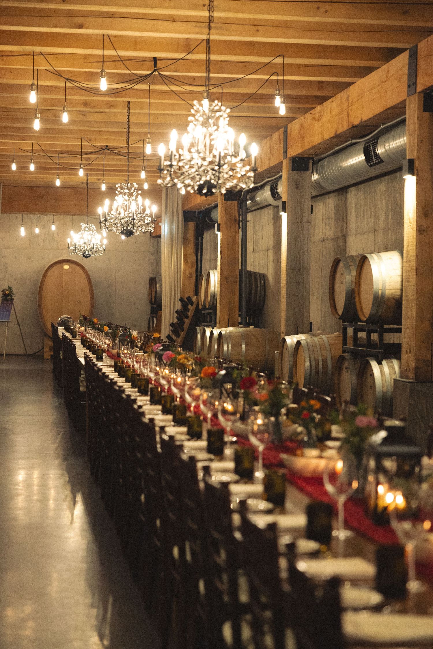 Long reception tables set under wooden beams and crystal chandeliers in rustic elegant venue.