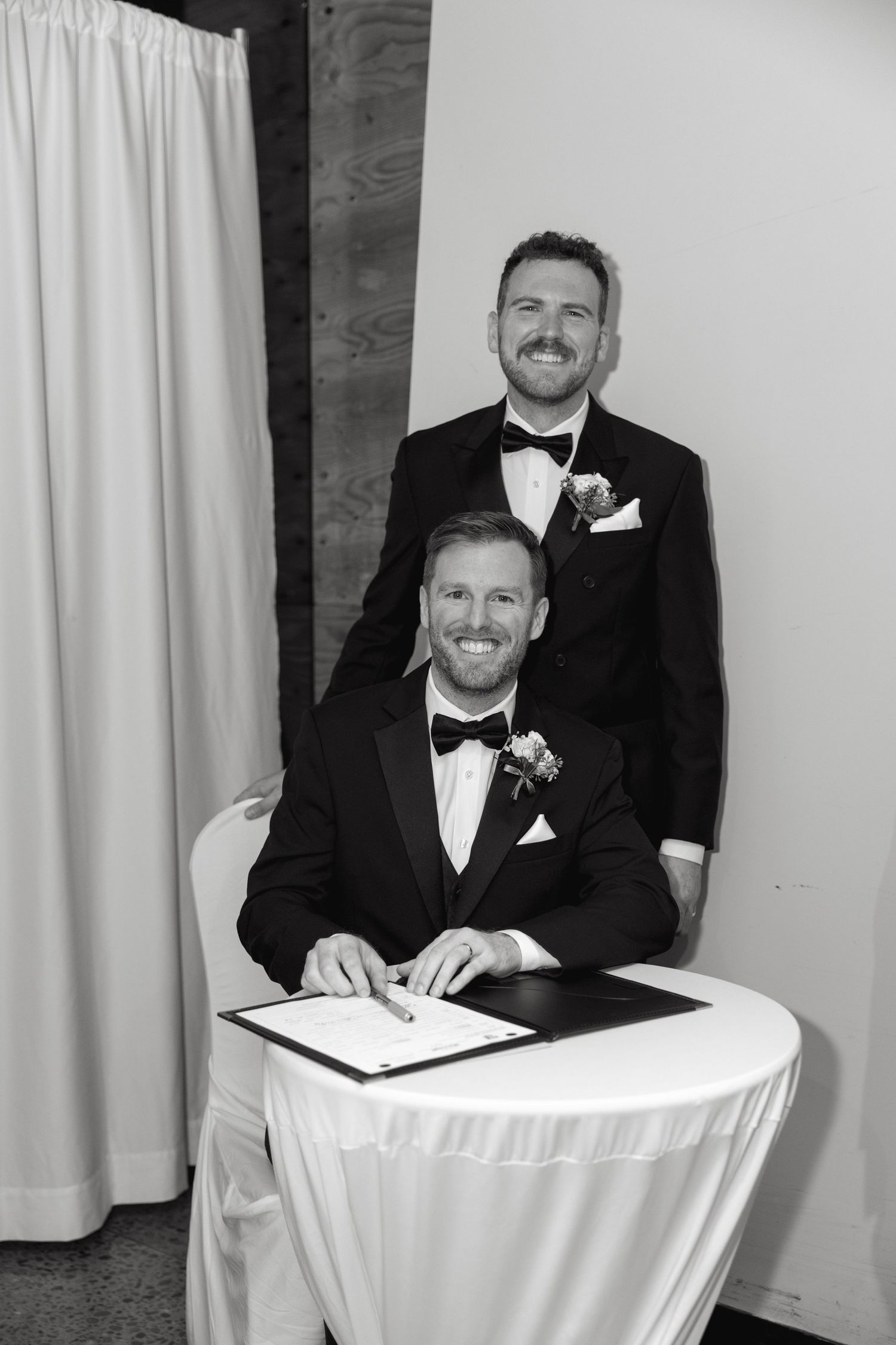 Groomsmen in black tuxedos and bow ties signing wedding documents at a white-clothed table.