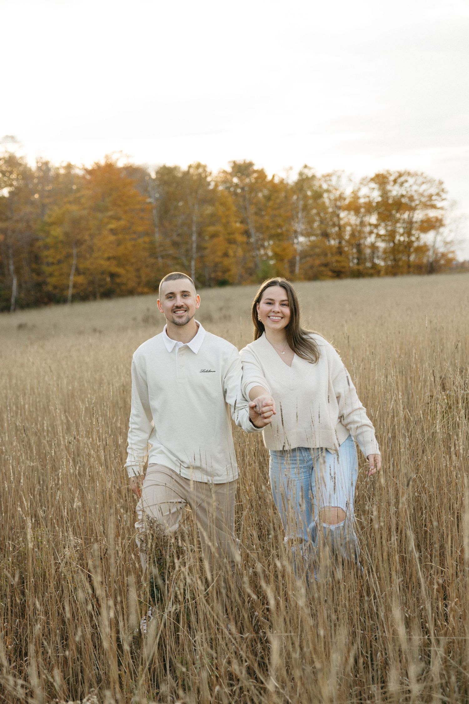 Happy couple walking through tall grass field during golden hour with fall foliage behind them.