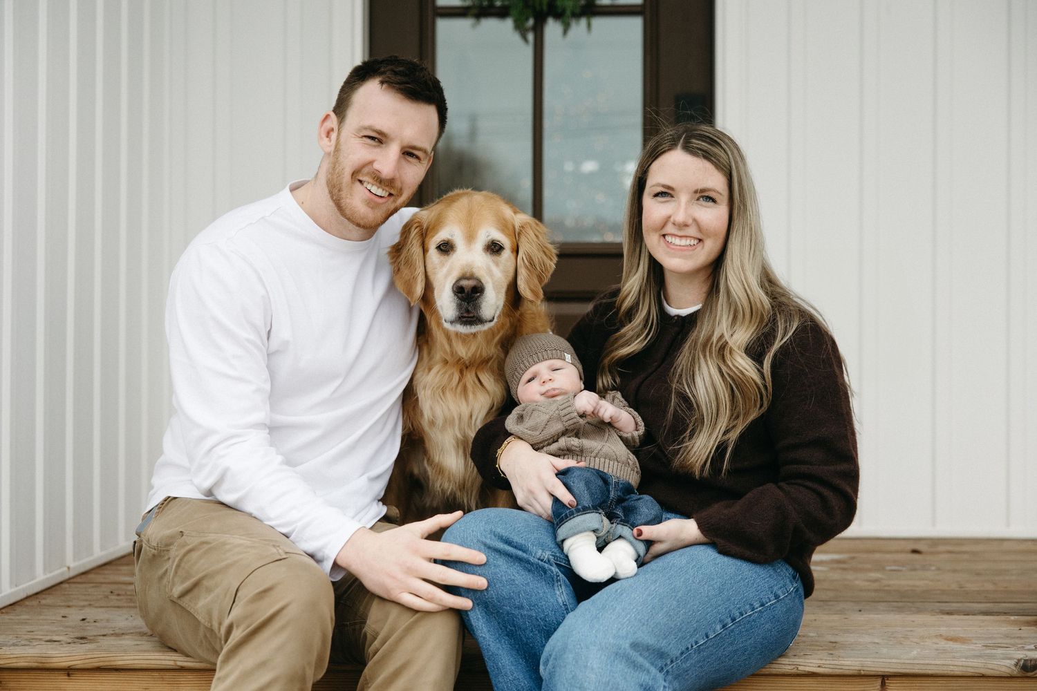 A couple sits on a wooden porch with their golden retriever during a holiday photo session.