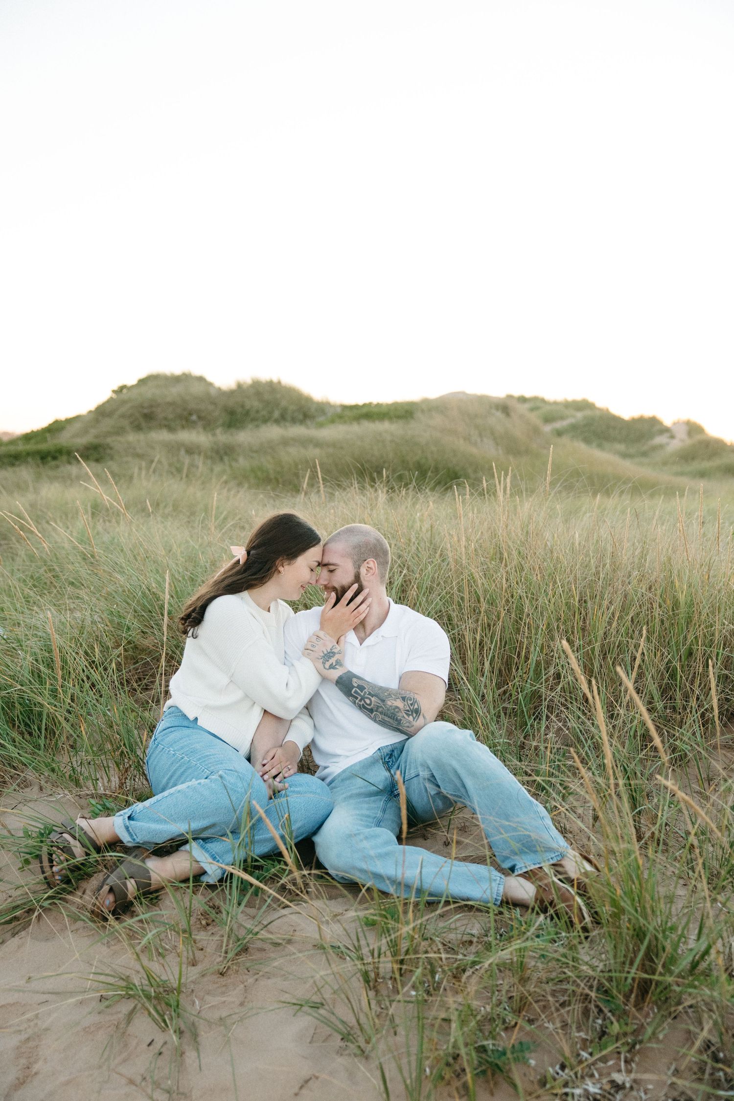 Couple sharing an intimate moment while resting on sandy dunes surrounded by beach grass.