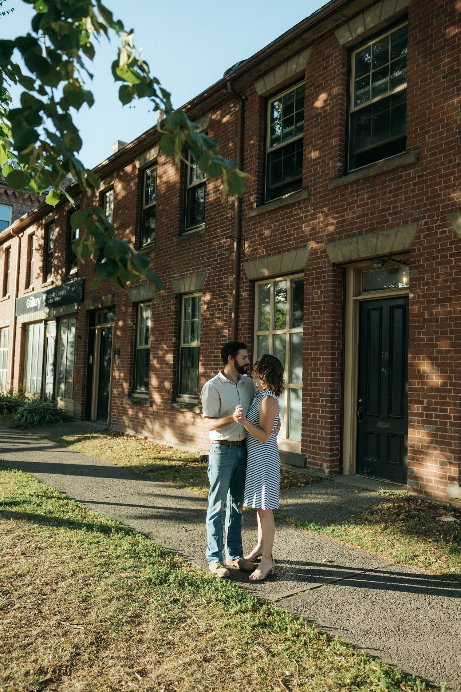A couple embraces on a sidewalk in front of a classic brick building with tall windows.