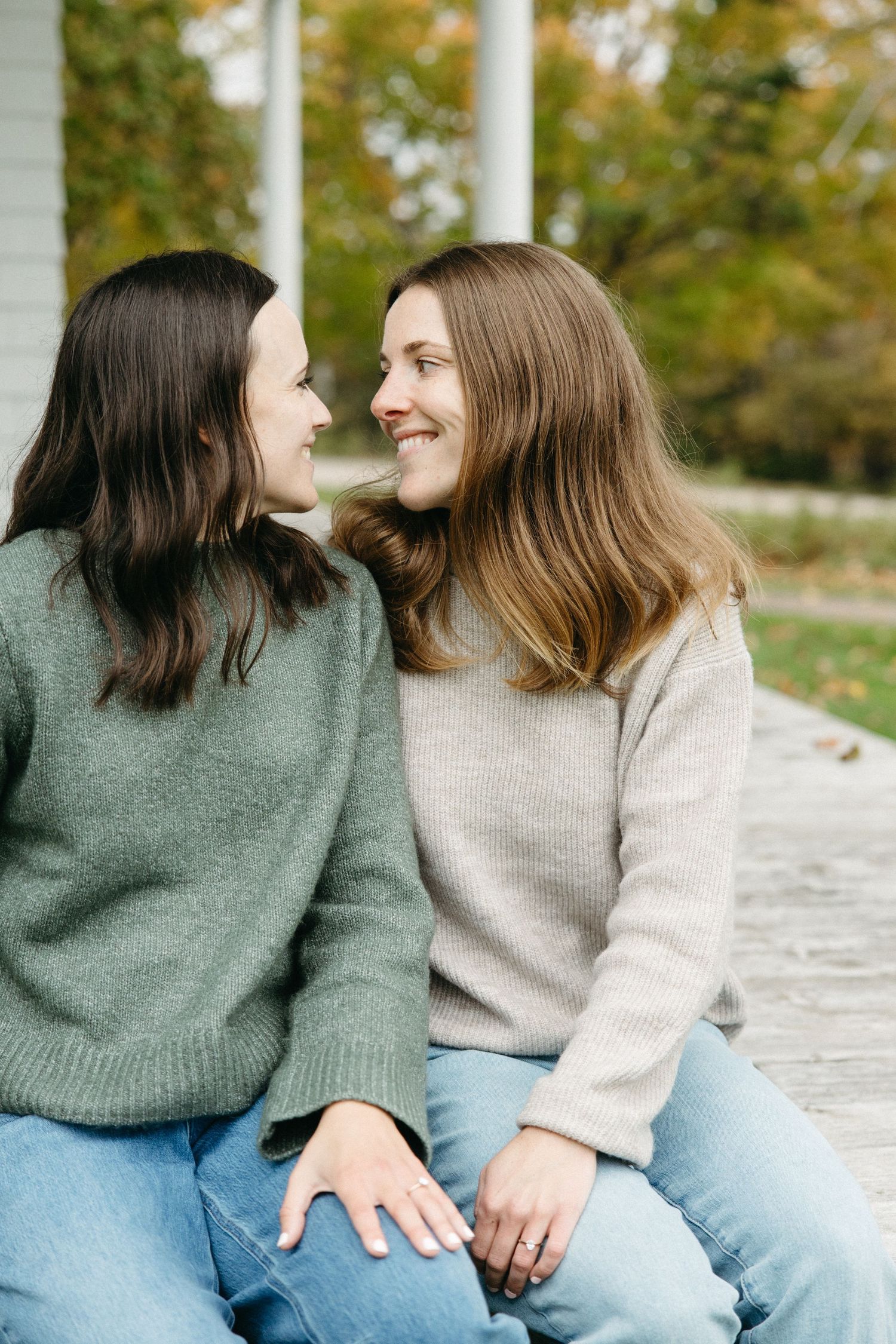 Two smiling women in cozy sweaters and jeans sit close together on a wooden deck, gazing warmly at each other with autumn trees behind them.