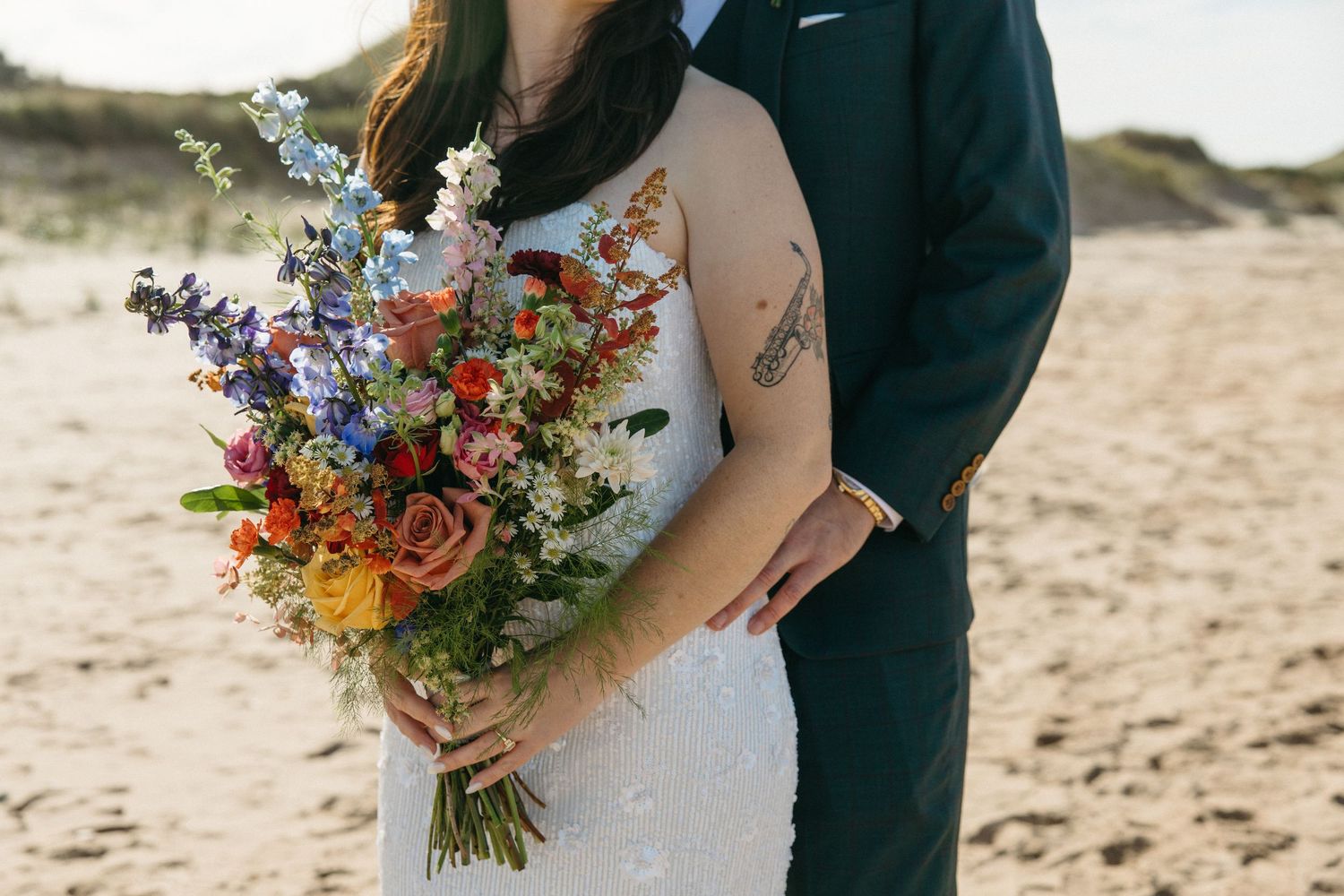 Bride in a white gown holds a colorful wildflower bouquet while her groom in a dark suit embraces her from behind on a sandy beach.