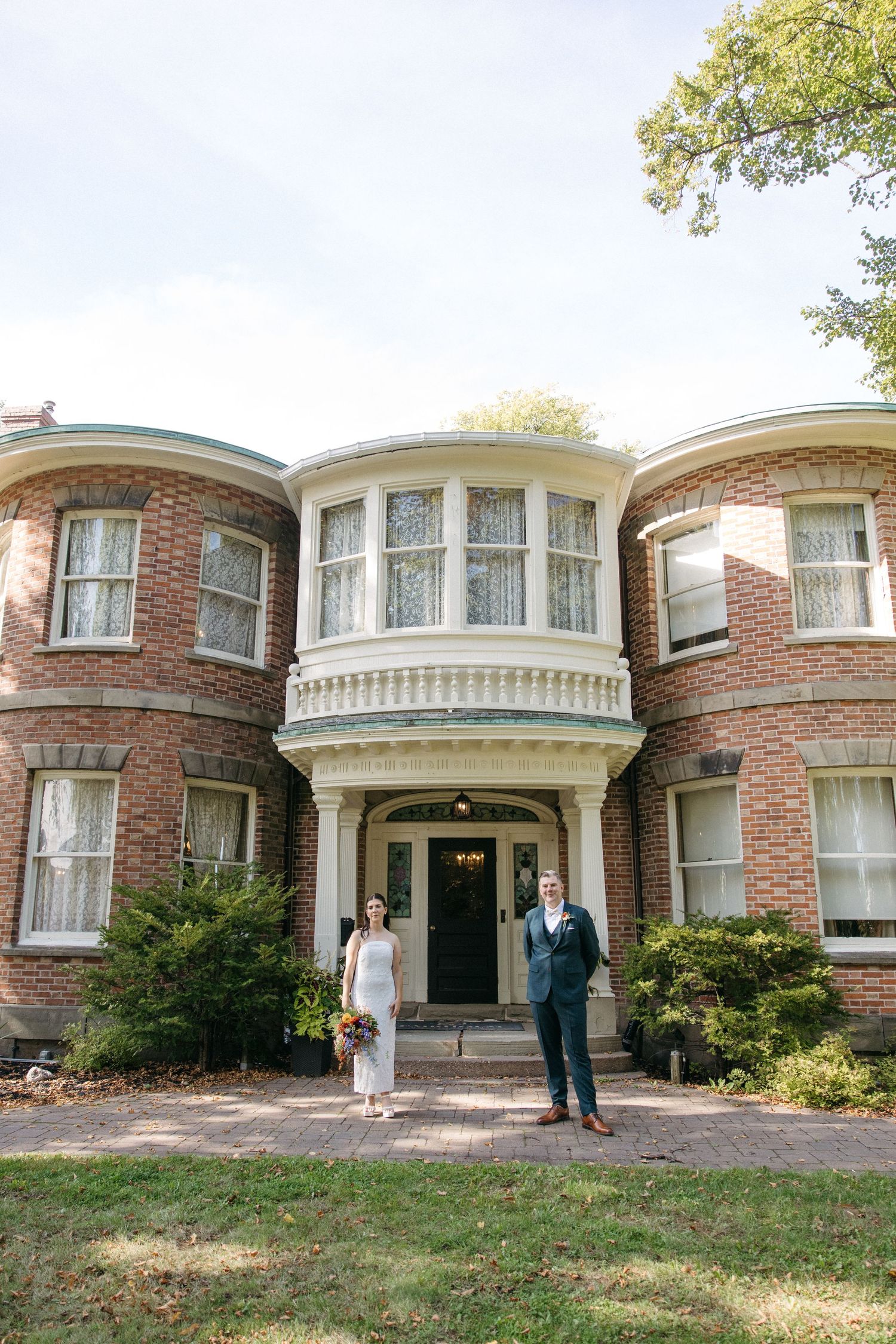 Bride in a white dress and groom in a blue suit stand before the entrance of a historic red brick mansion with a bay window.