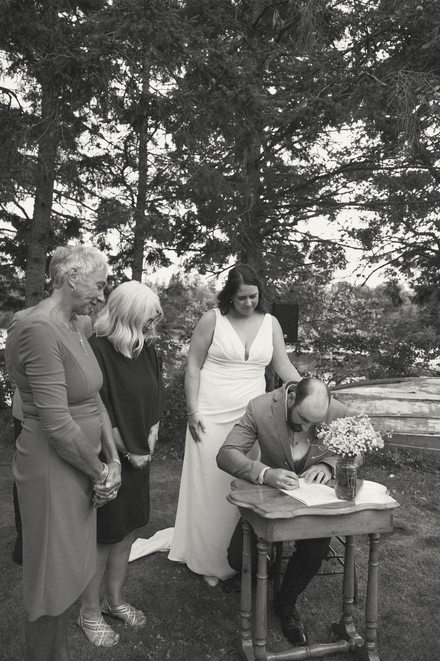 Wedding ceremony guests signing documents at outdoor table under trees.
