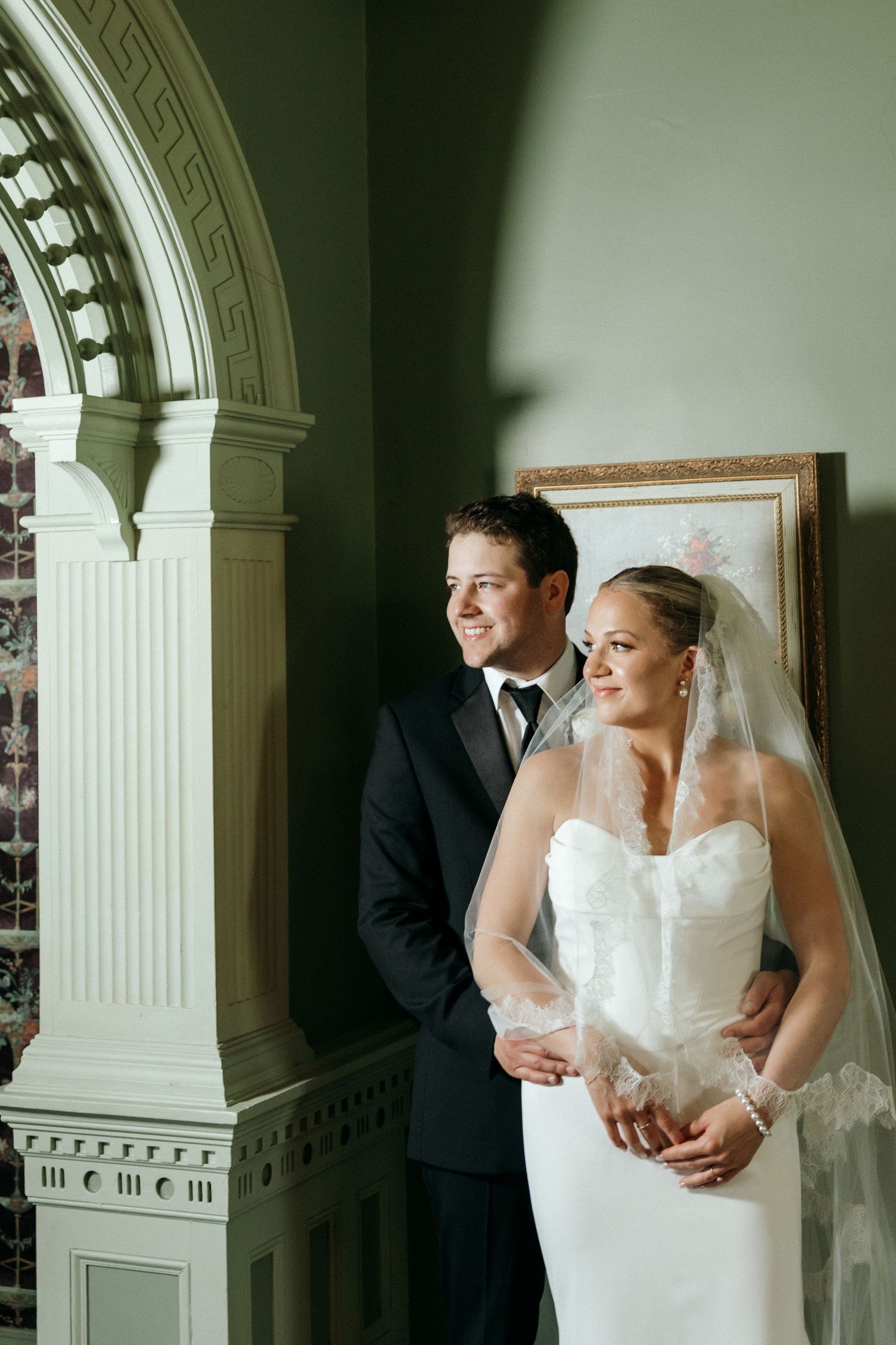 A wedding couple poses together against a white column in an elegant interior setting.