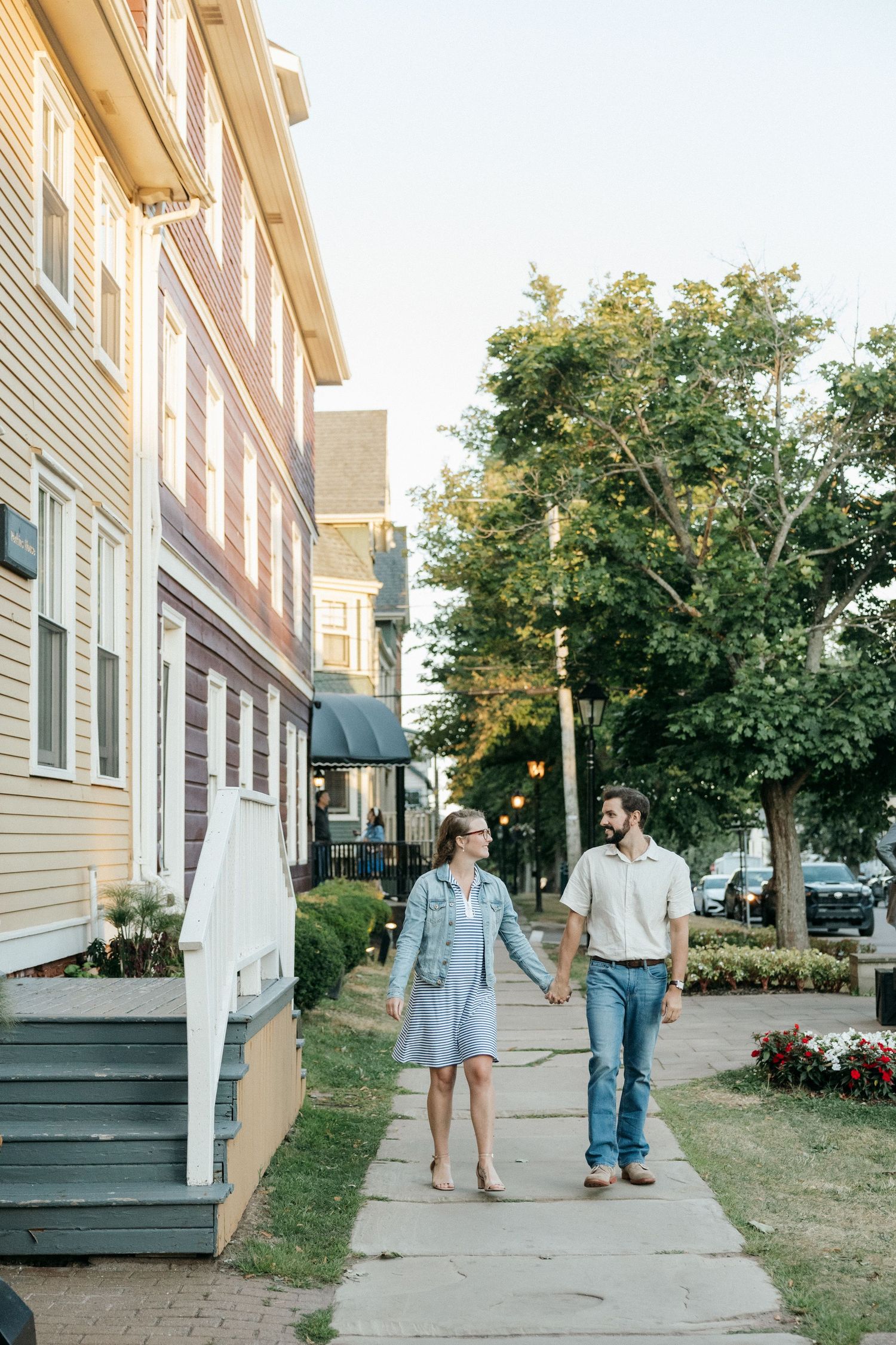 Two people walking along residential sidewalk past historic houses in evening light.