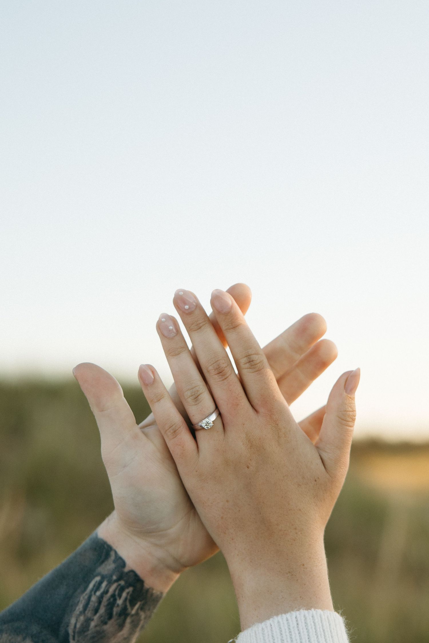 Close up of hands with engagement ring intertwined against bright sky background.