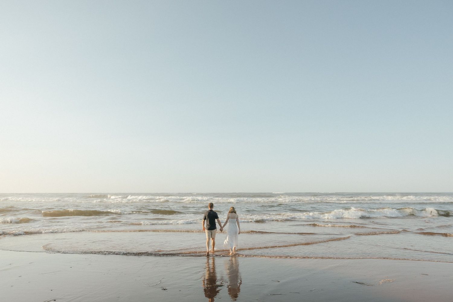 A couple walks along a misty beach shoreline with waves crashing in the background during golden hour.