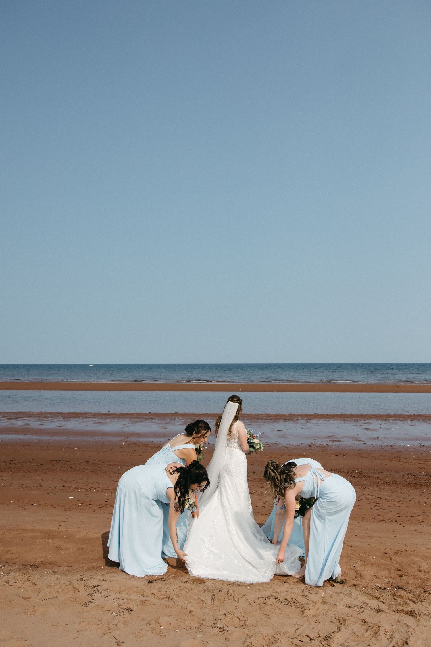 Wedding party members in light blue dresses assist with a white wedding gown on a beach.