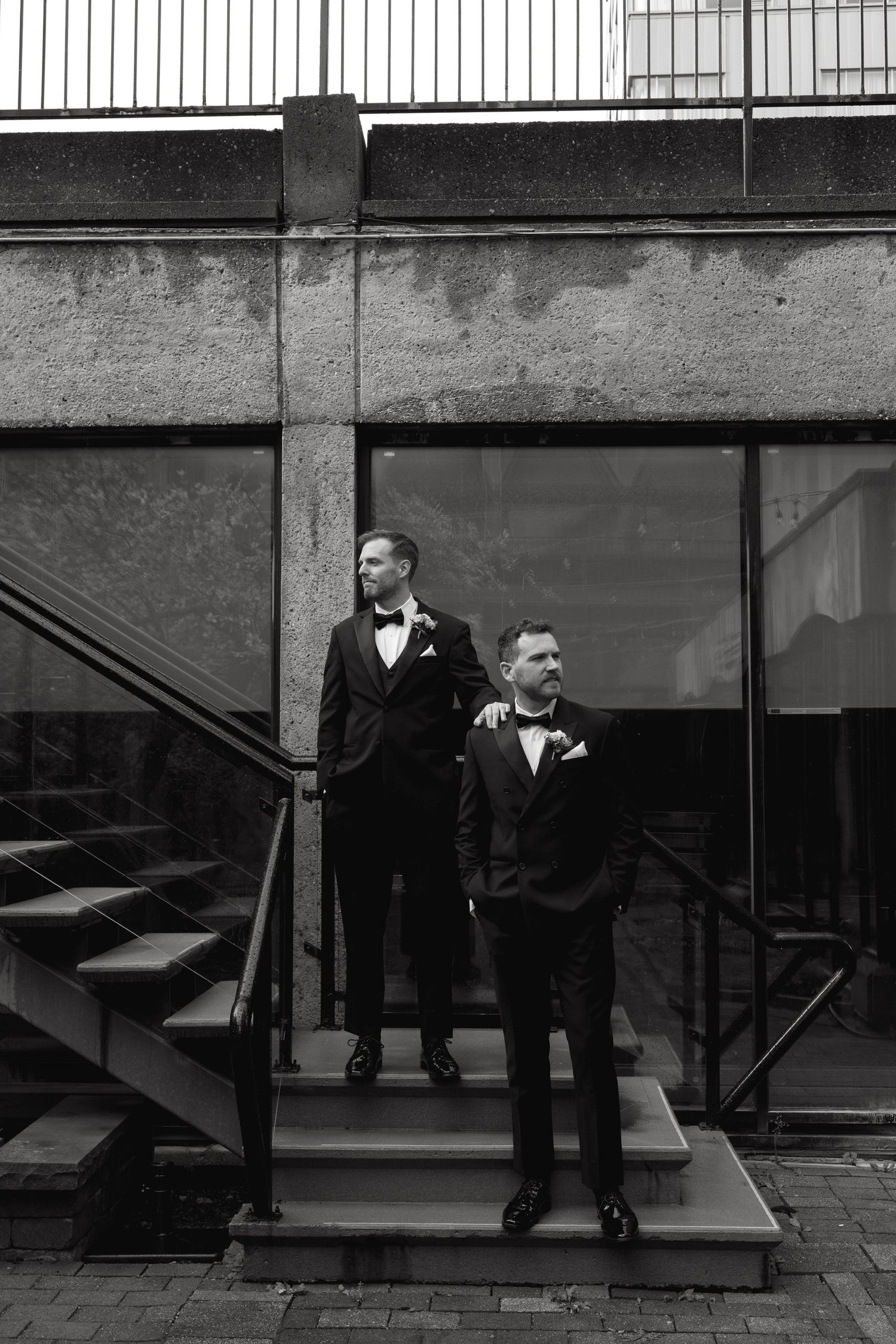 Two people in formal black tuxedos stand on concrete steps against an industrial backdrop.