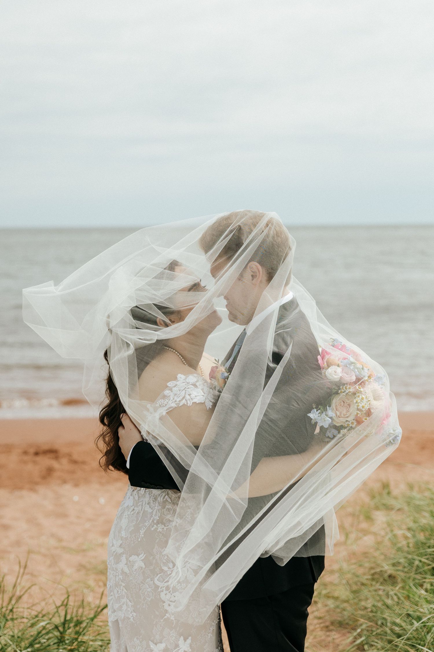 A wedding couple embraces on a beach as the veil flows in the ocean breeze.