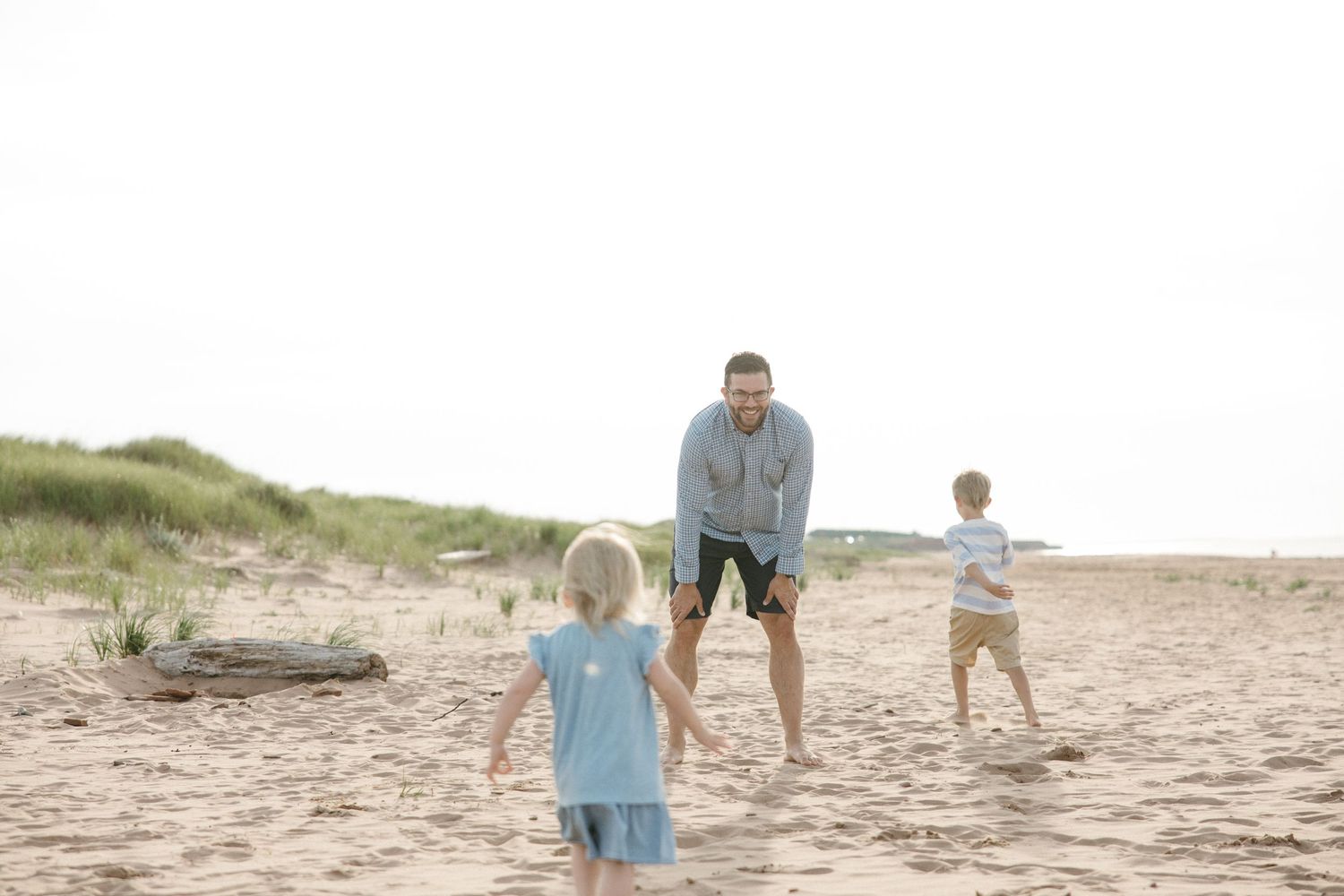 A father plays with his two young children on a sandy beach with grass dunes in the background.