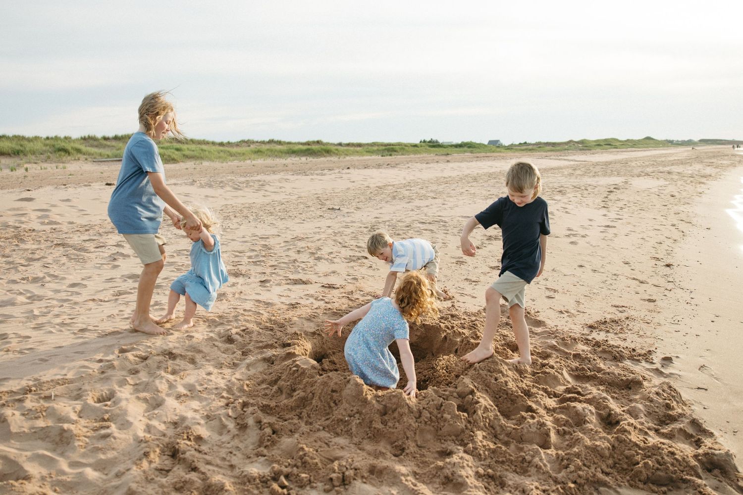 Four children building a sandcastle together on a wide sandy beach on a cloudy day.