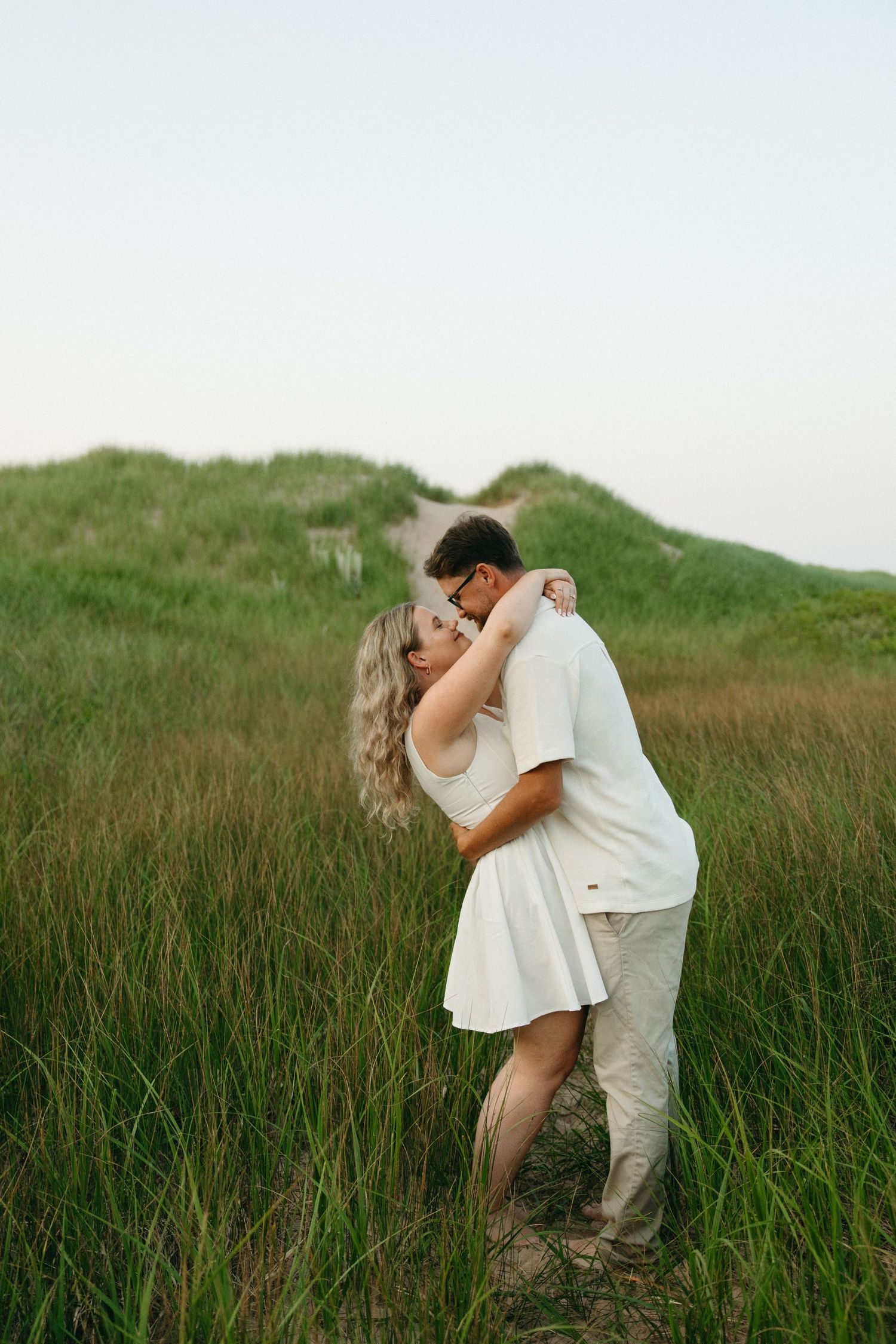 Romantic couple embracing and kissing in lush green coastal grass dunes at sunset.