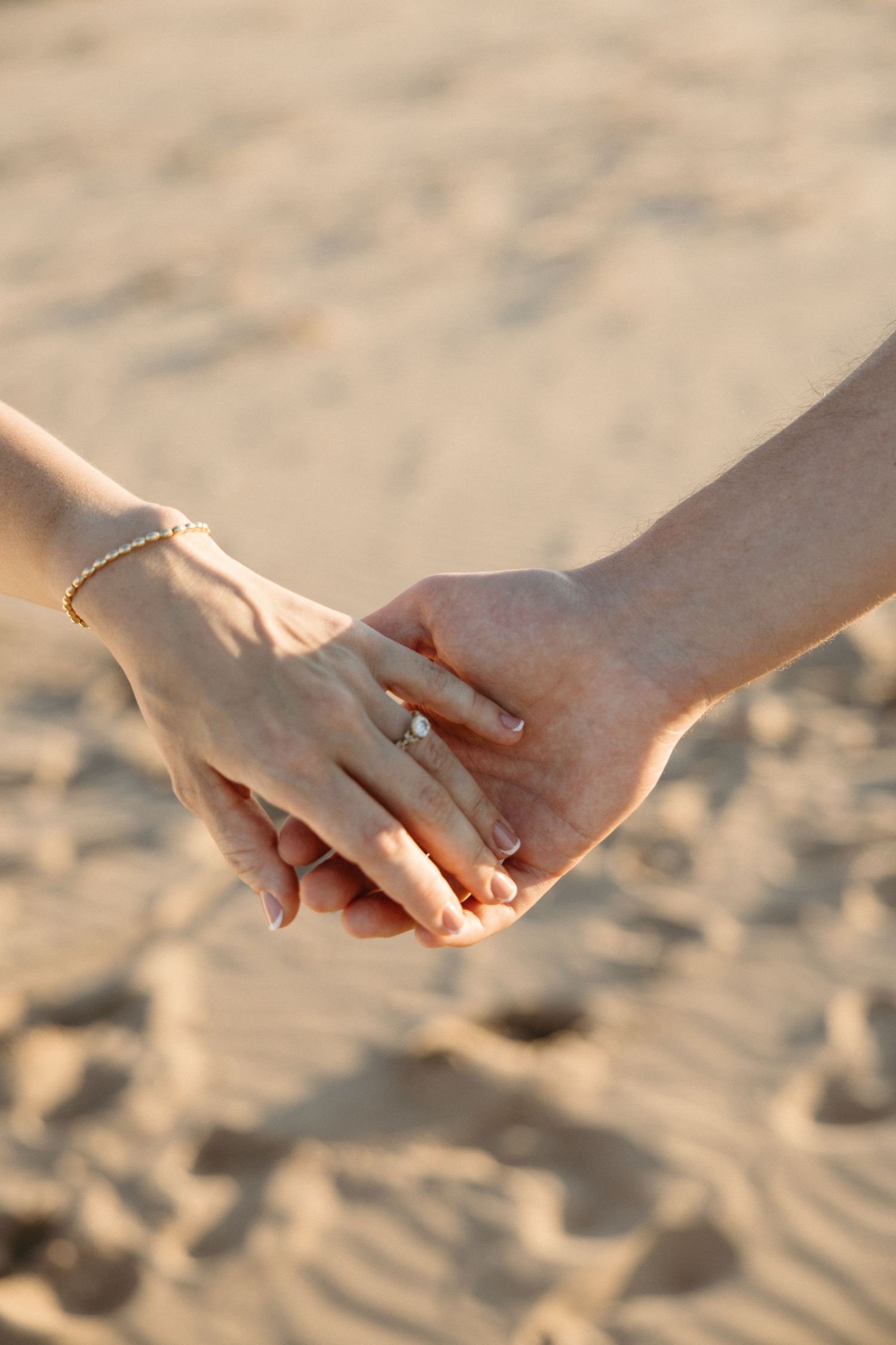 Close-up of couple holding hands on sandy beach, woman wearing diamond engagement ring and gold bracelet.