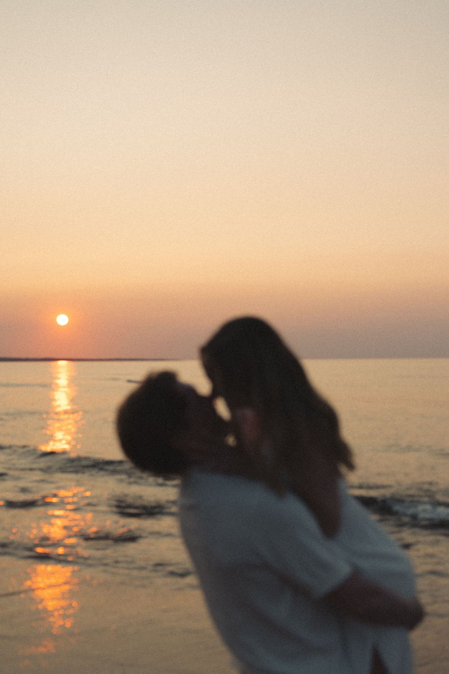 Couple sharing a kiss silhouetted against a glowing orange sunset reflecting on calm ocean water.