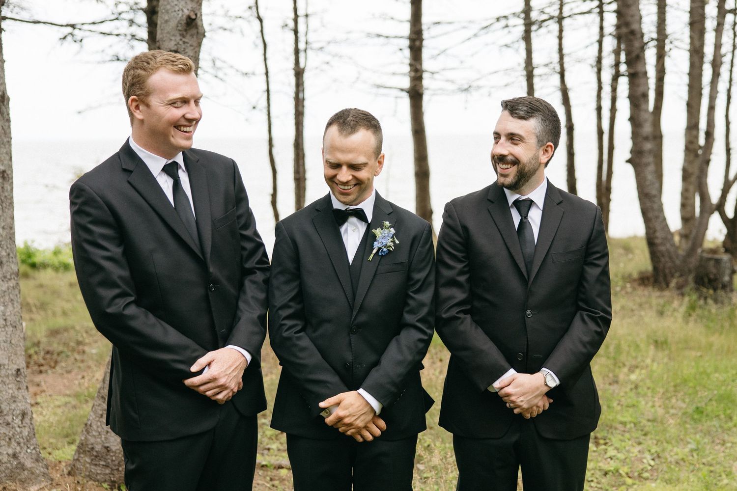 Three groomsmen in black suits smile together outdoors among trees at a wedding celebration.