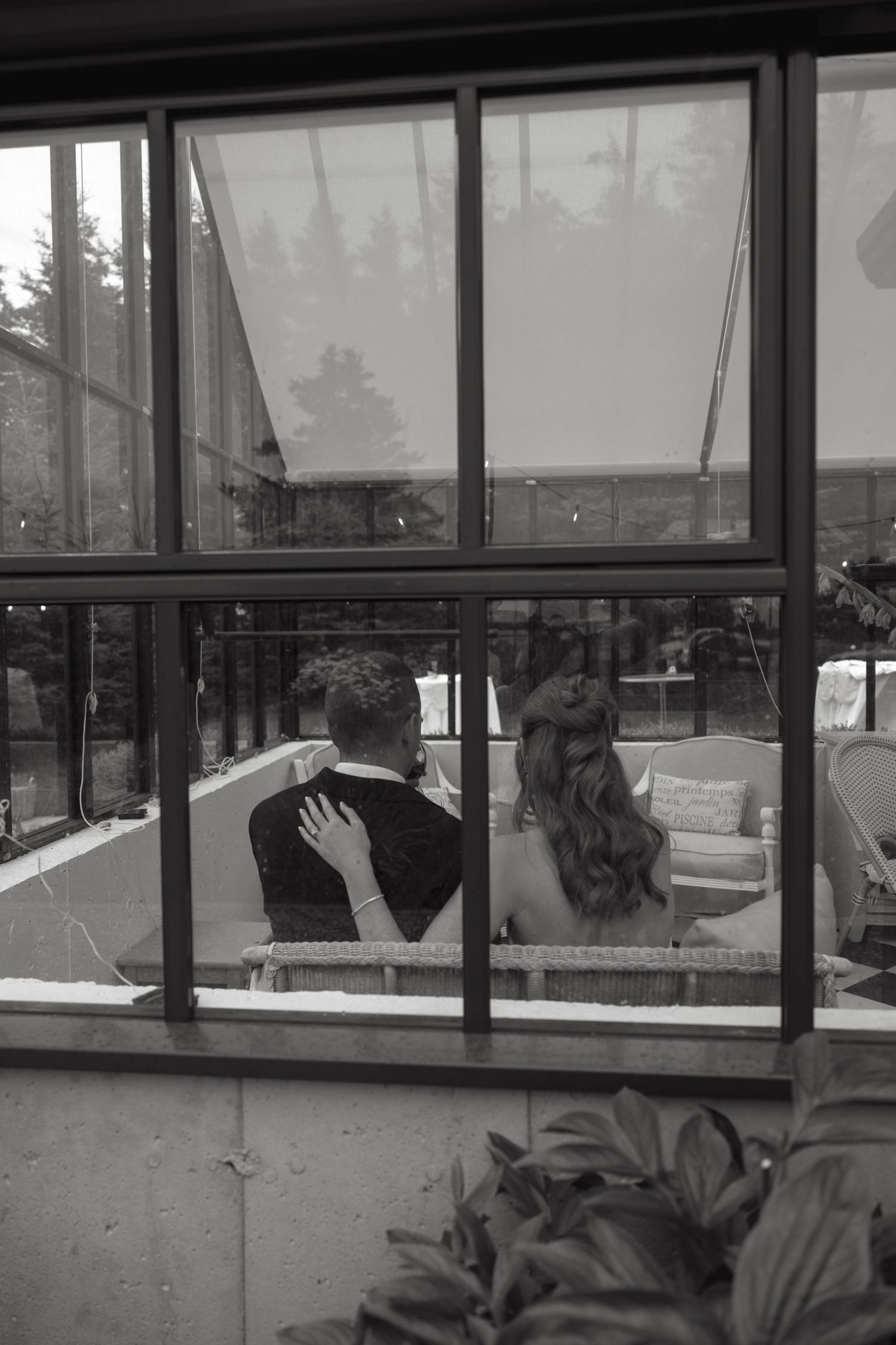 Black and white photo of bride and groom sitting together viewed through large glass windows from outside.