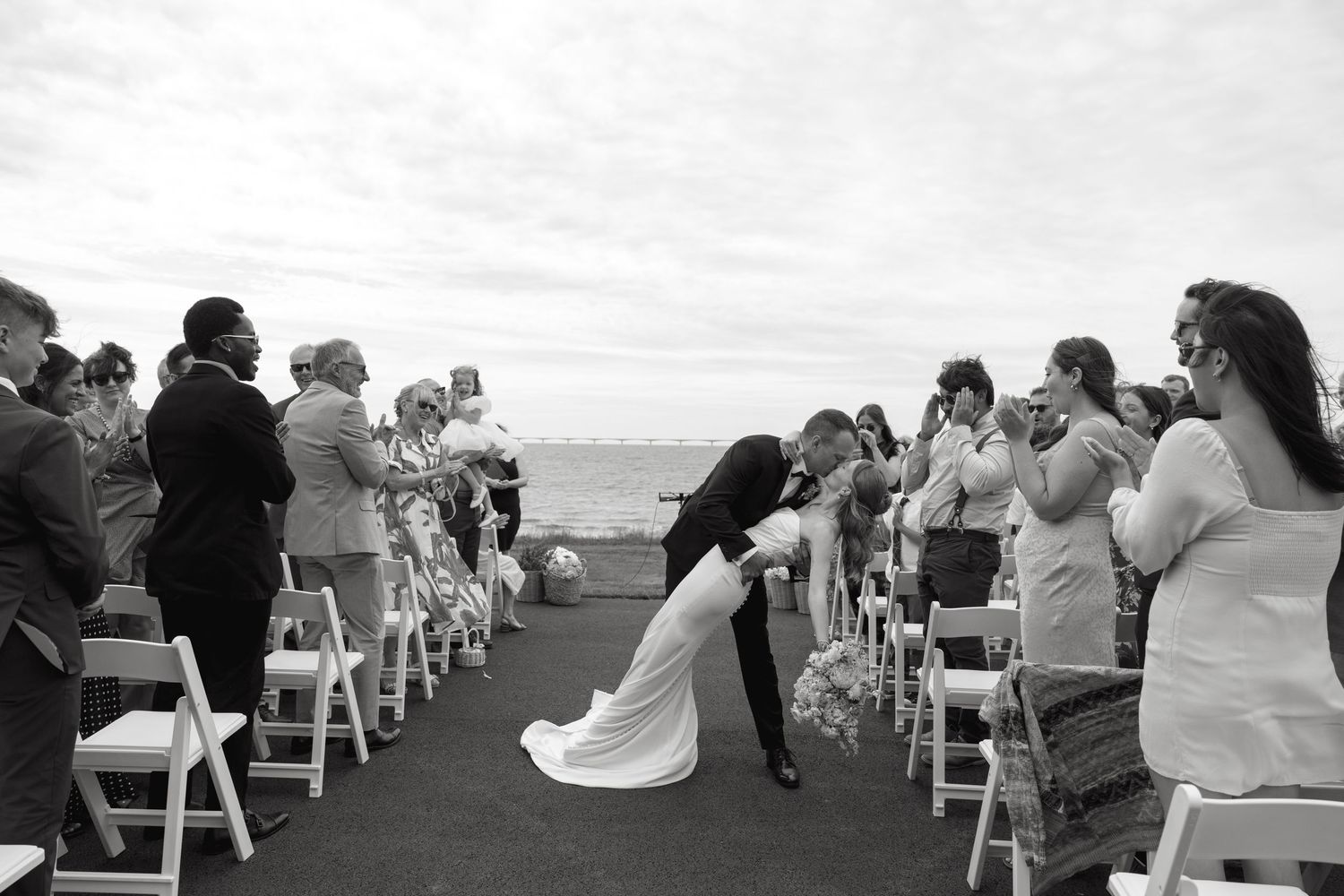 Black and white photo of groom dipping bride in wedding aisle surrounded by standing applauding guests outdoors.