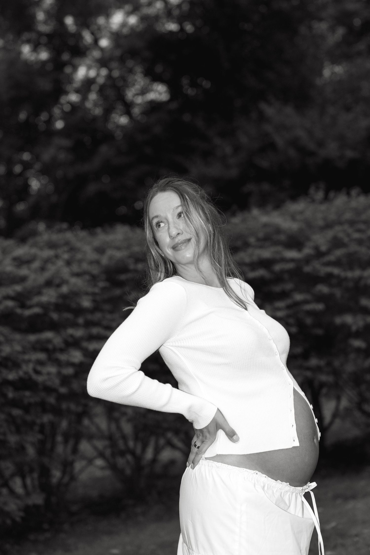 Black and white portrait of pregnant woman in white outfit posing in garden, showing baby bump.