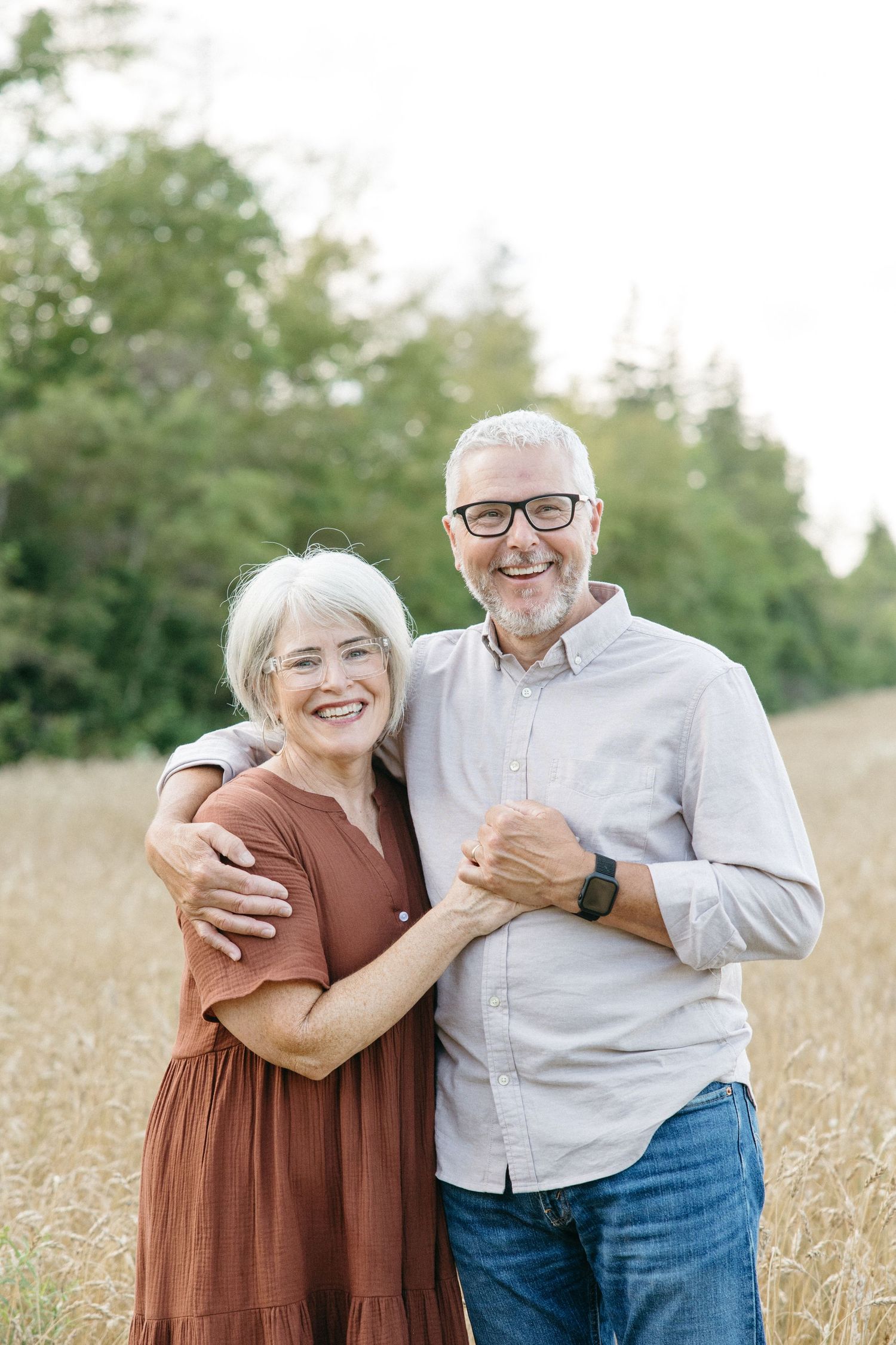 Smiling older couple embracing in a golden field, woman in rust dress, man in gray shirt and glasses.